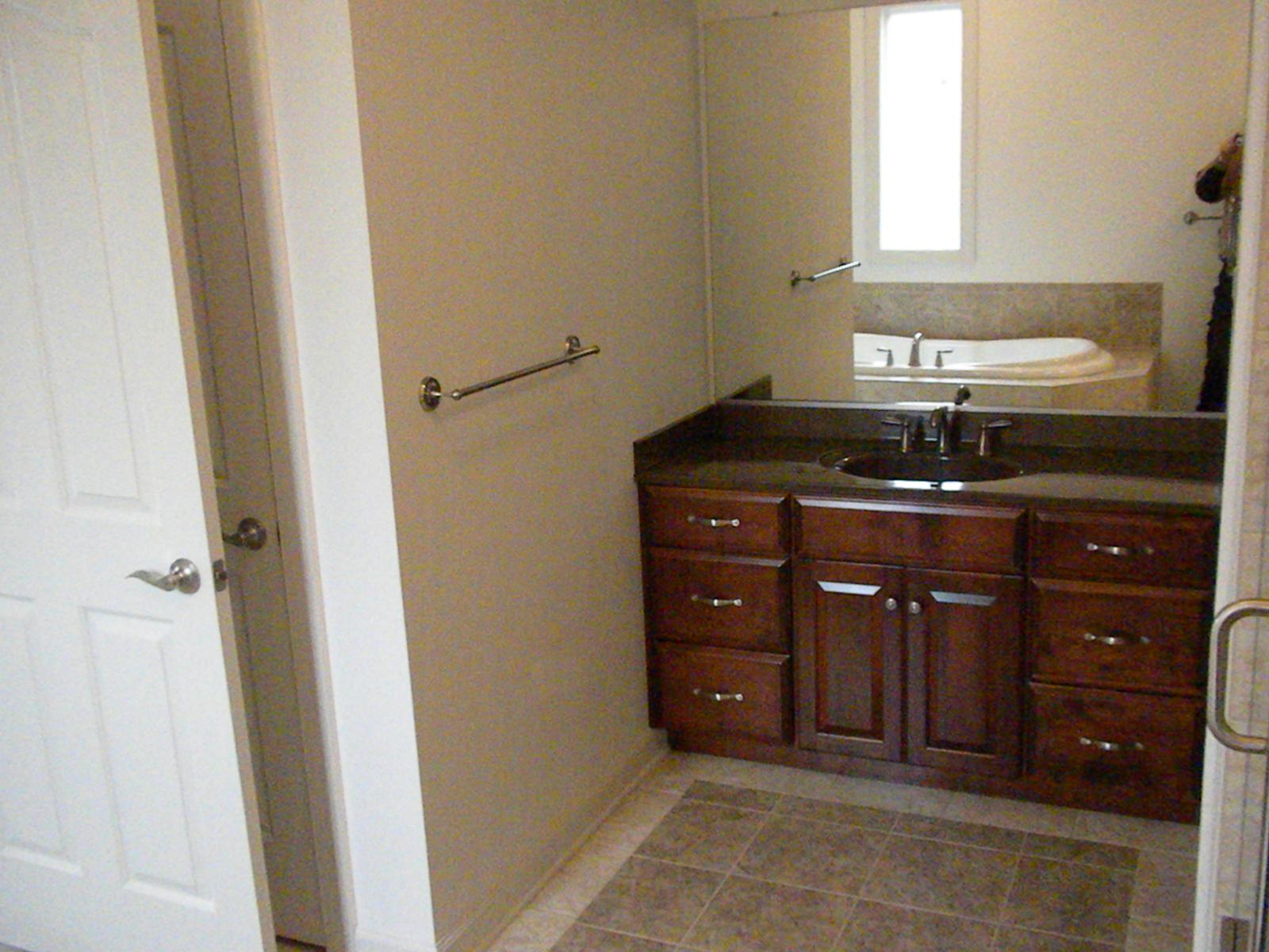 A bathroom vanity with a stone top and dark cabinets, a tall narrow window above and a door standing open