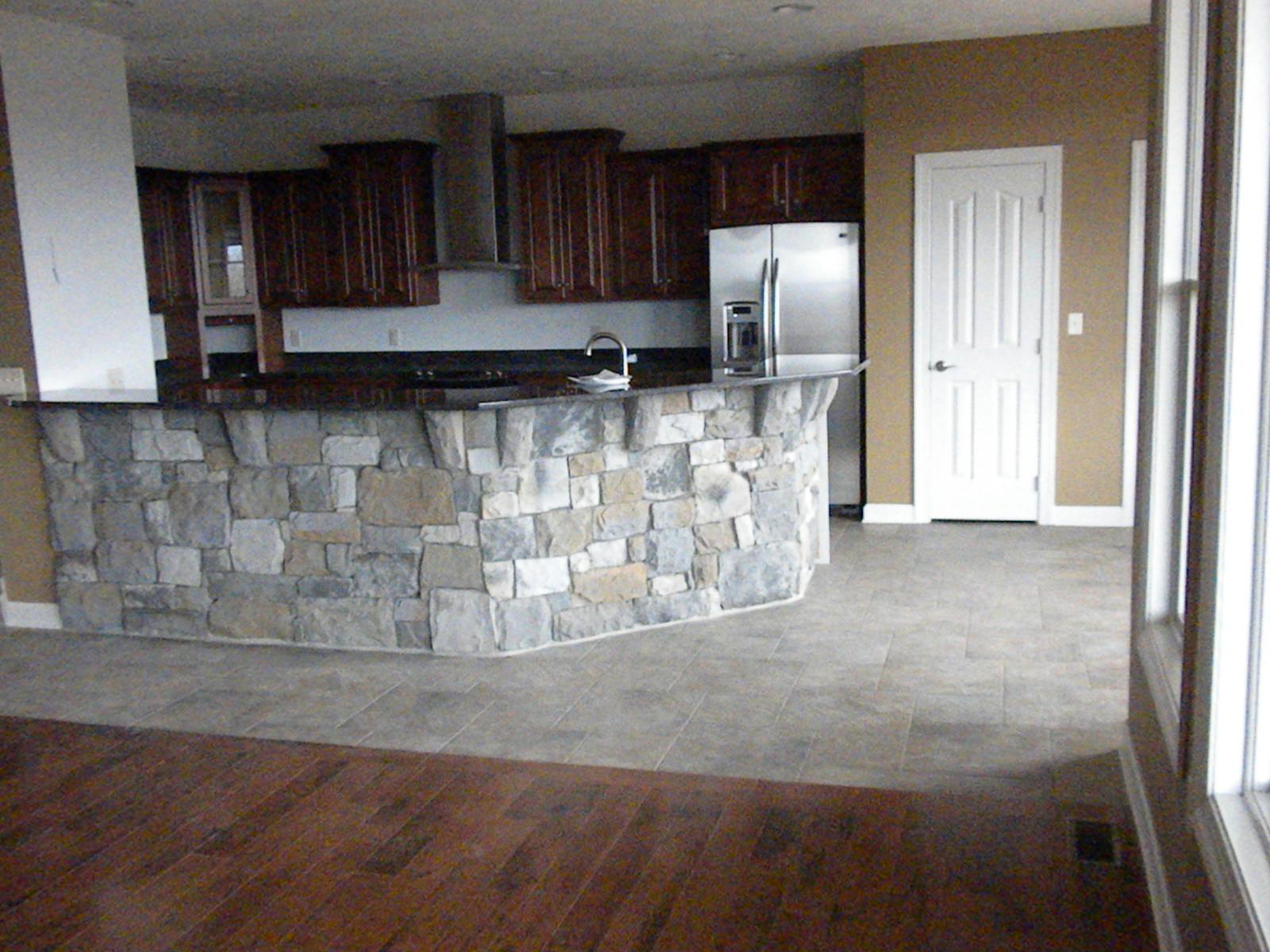 A kitchen behind a curved stone-faced bar, dark cabinets and a stainless refrigerator along the far wall