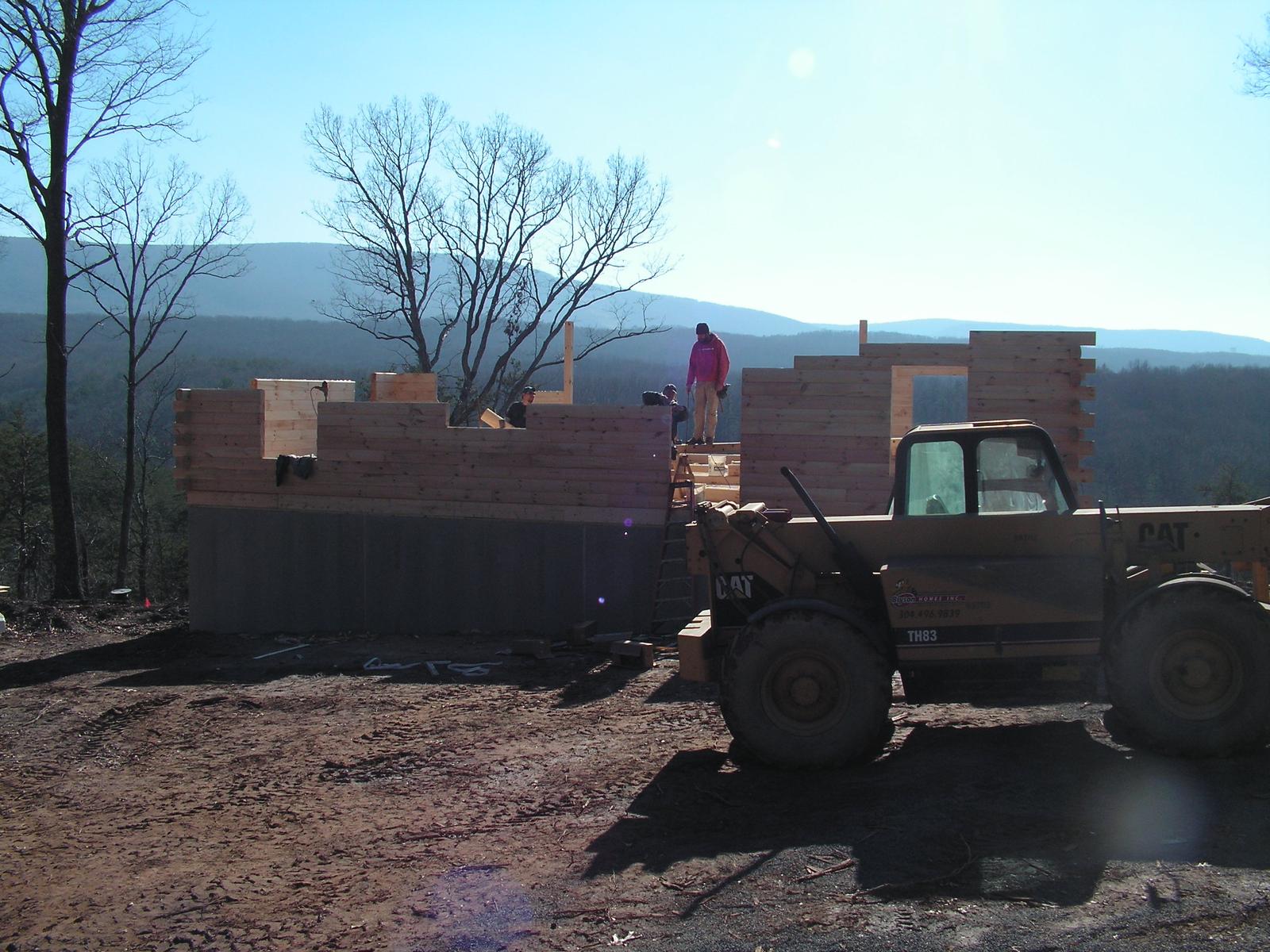 Two people working on top of the log walls as a telehandler stands by, mountains behind