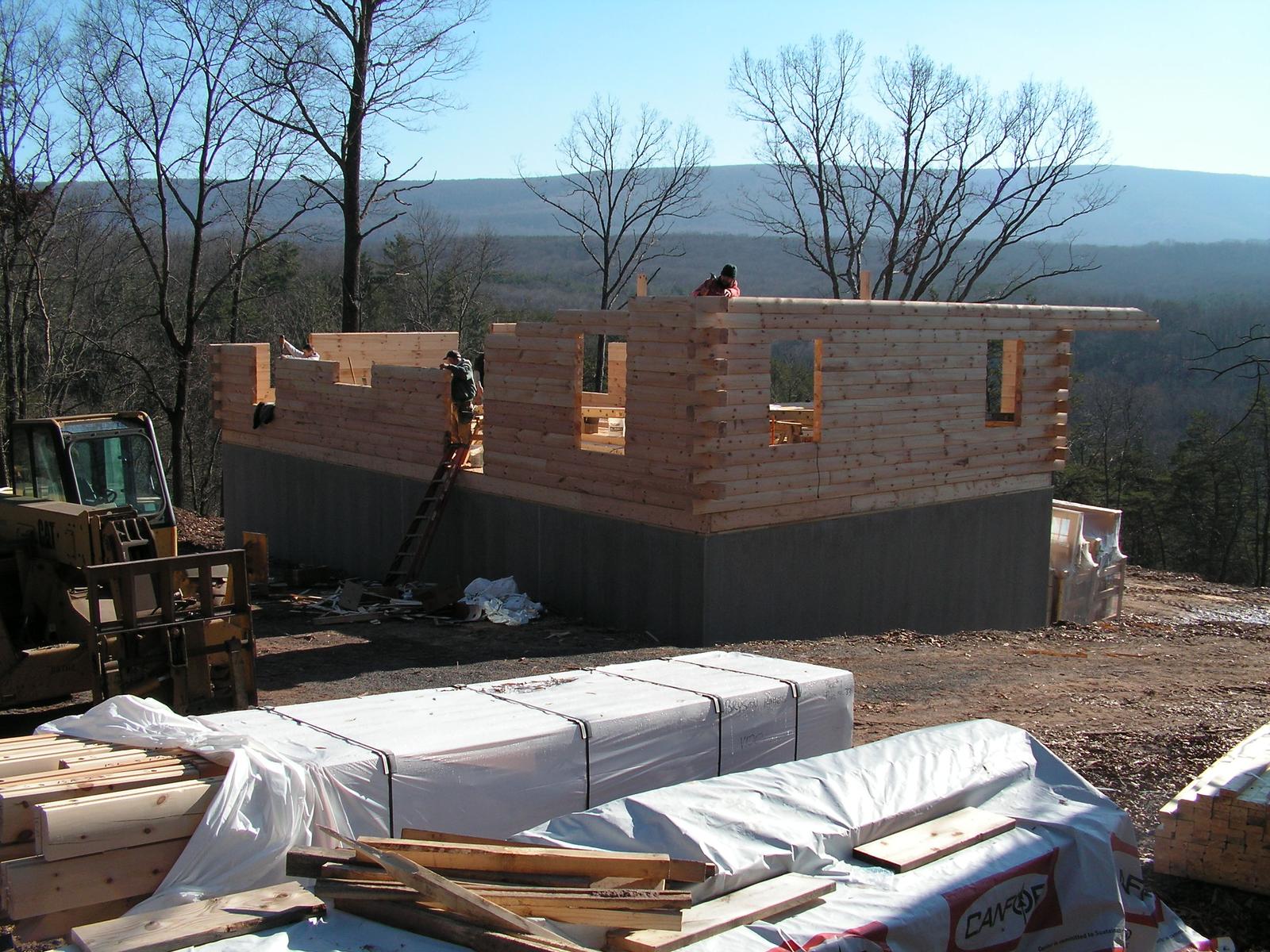 Log walls part stacked on a dark foundation, packaged logs on pallets in the foreground and a ridge beyond