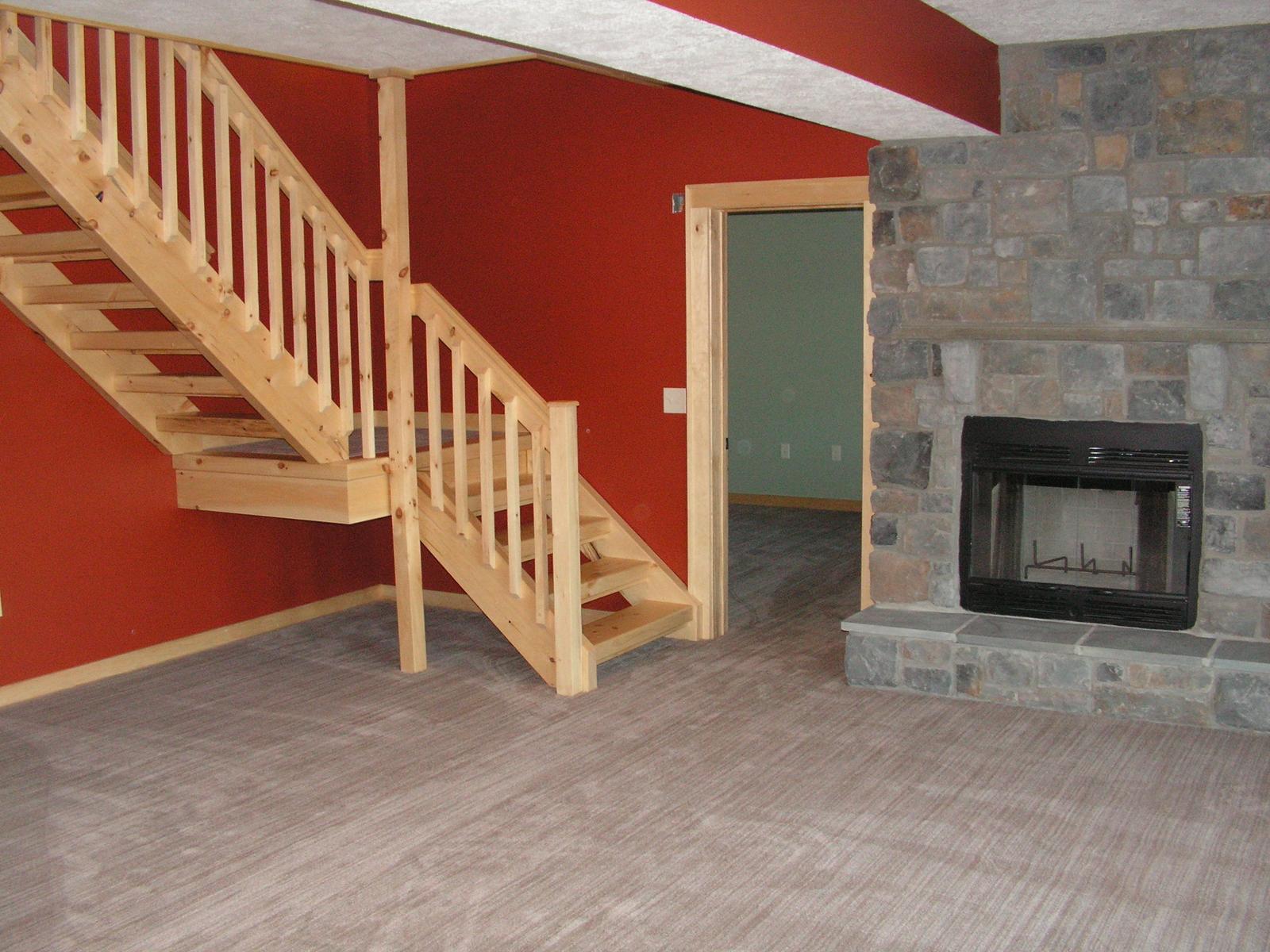 A stair running down beside a stone chimney into a lower-level room, red wall behind the treads