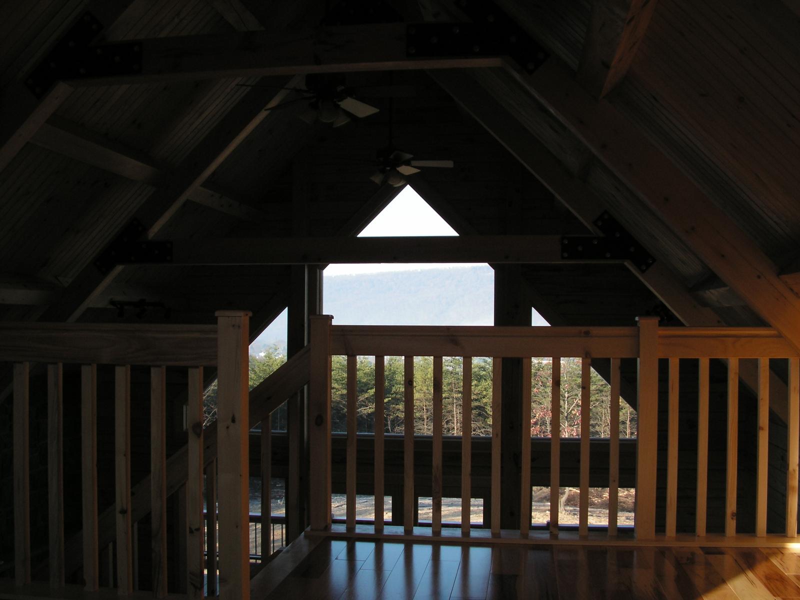 A loft in shadow with a triangular gable window at the ridge, timber rafters and a rail across the opening