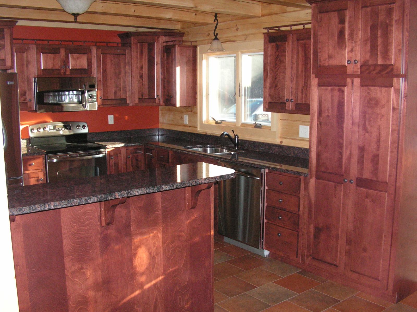 A kitchen with red-stained cabinets and dark stone counters, stainless range and dishwasher along the run