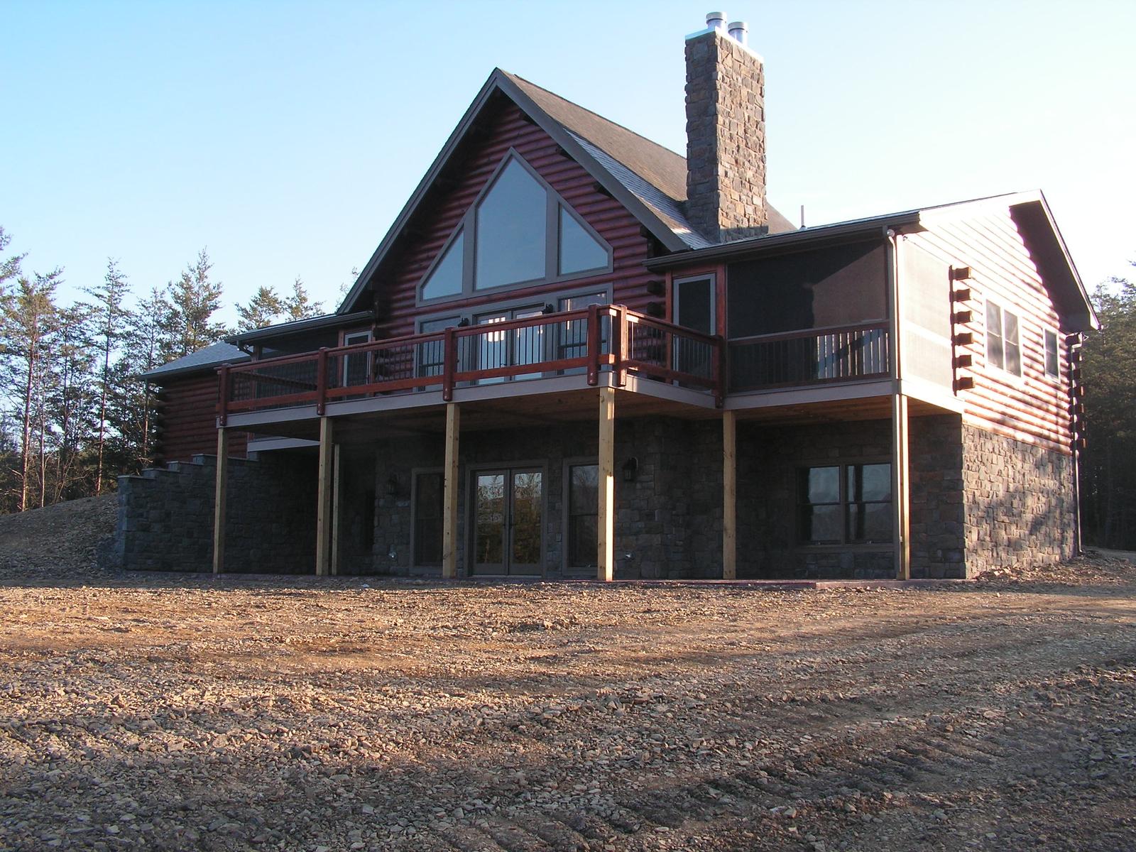 A log home with a gable of glass and a stone chimney, its deck carried on posts above a walkout level