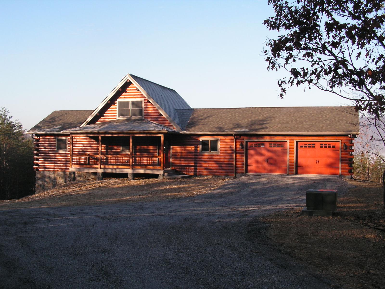 A long log home with a covered porch and an attached garage, red-stained walls under a low roof on a graded site