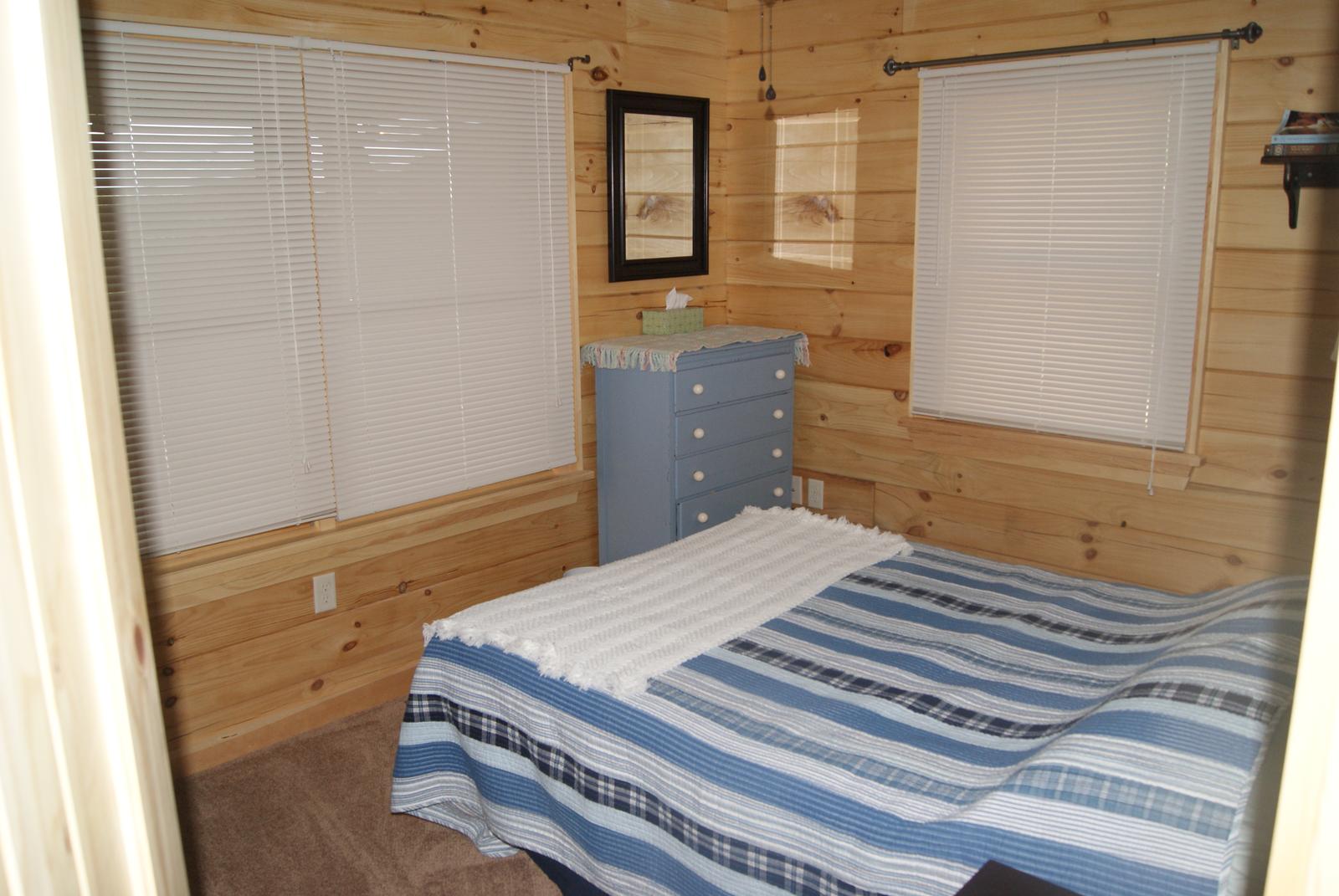 A bedroom with board walls and two shuttered windows, a striped cover on the bed