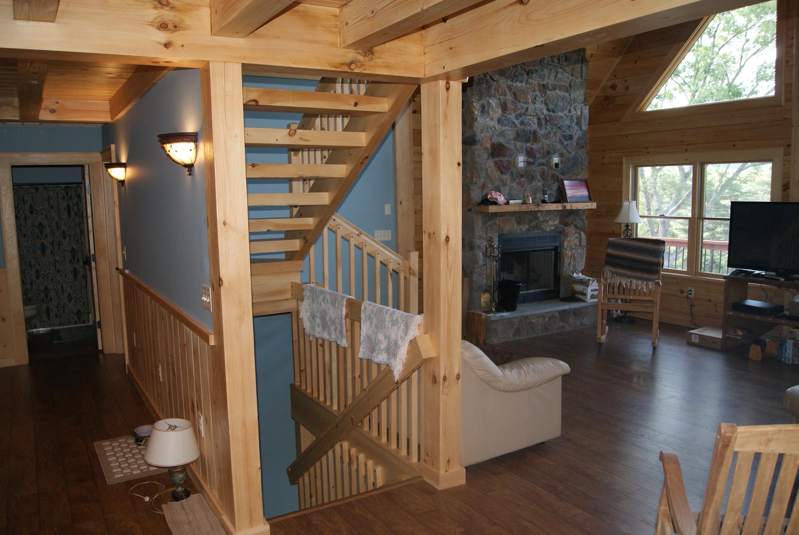 A stair with an open rail beside a stone chimney, the great room and its windows opening past it