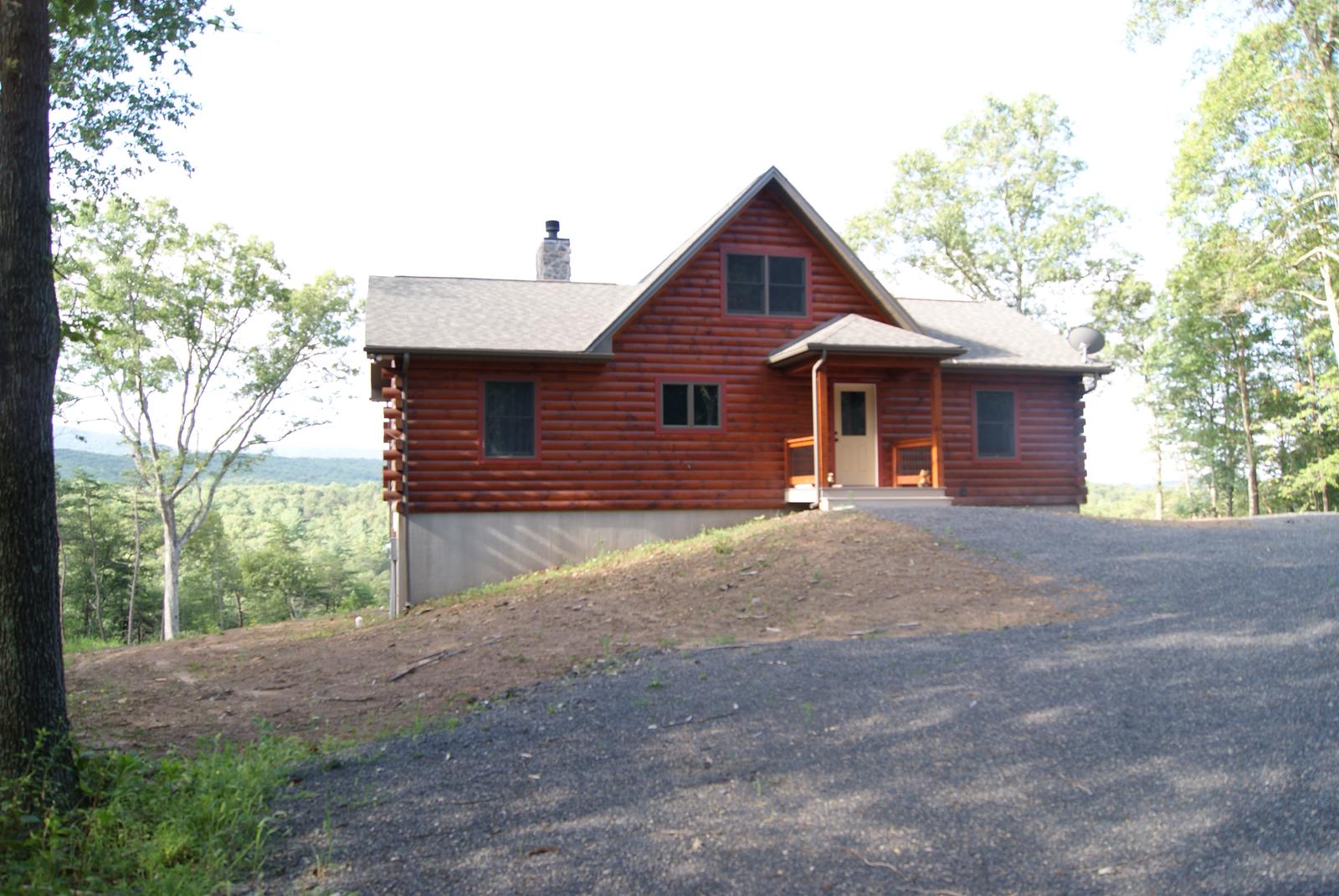 A log home on a graded pad with a gravel turning circle in front, a stone chimney at the ridge and green woods close on every side