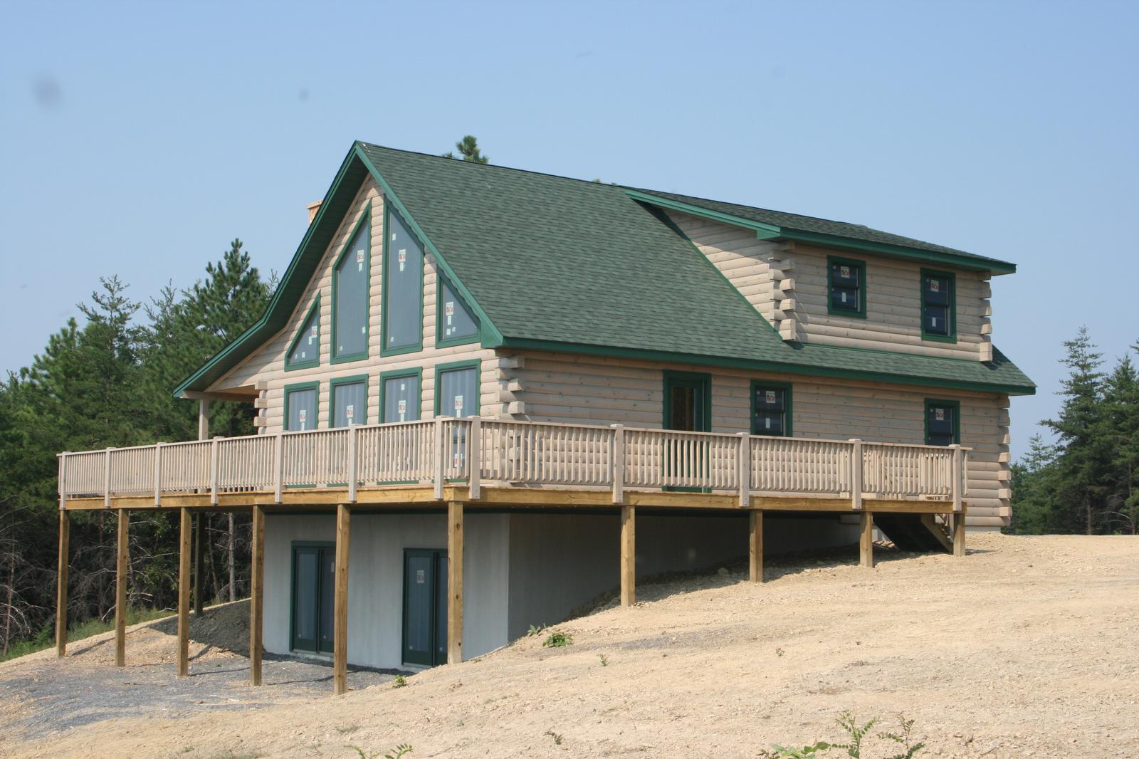 A log home with a green roof and a gable of glass, its deck carried on posts above a walkout level