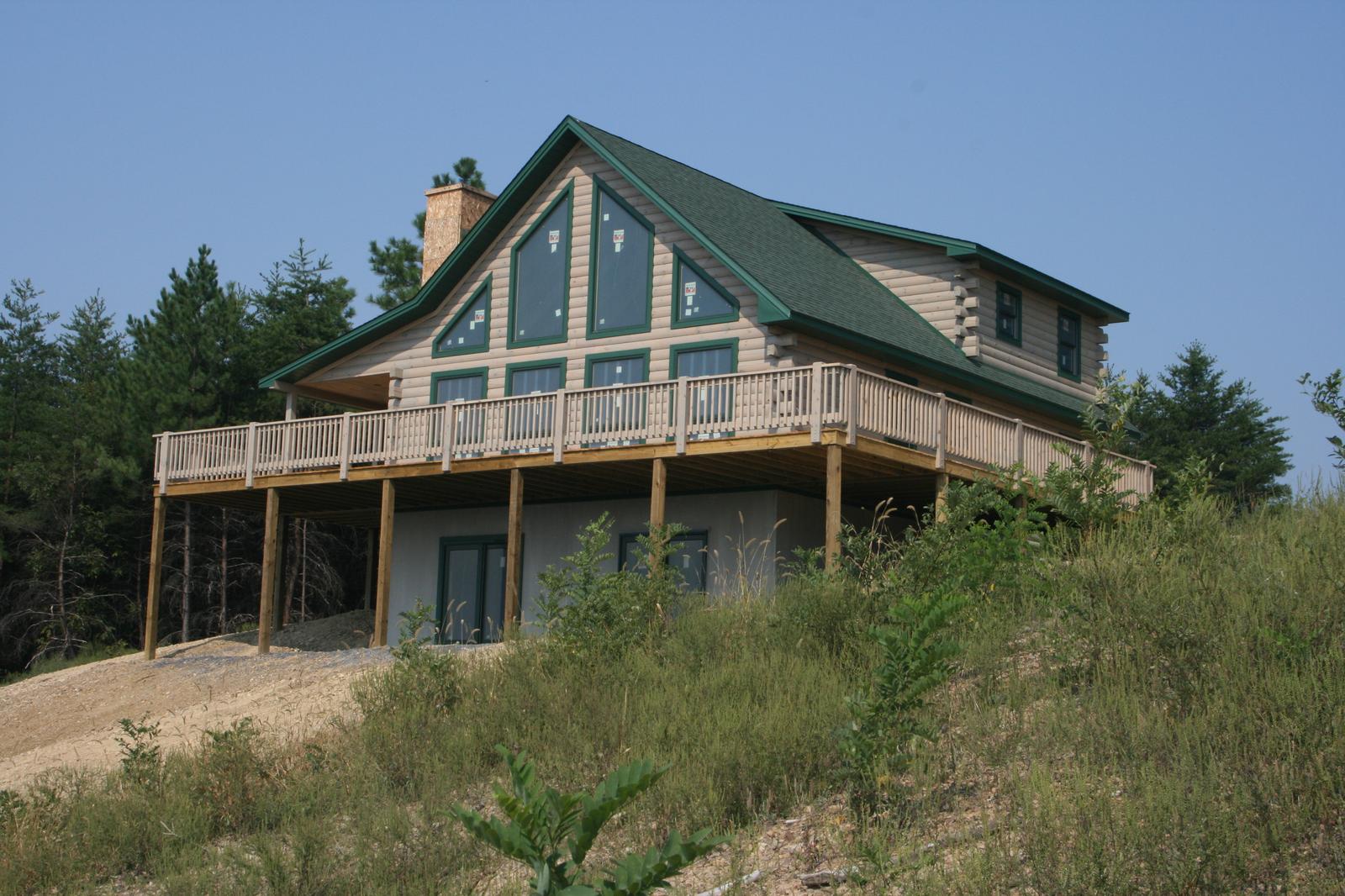 The home from below the bank, gable glass and green roof above the deck, scrub grown up in front
