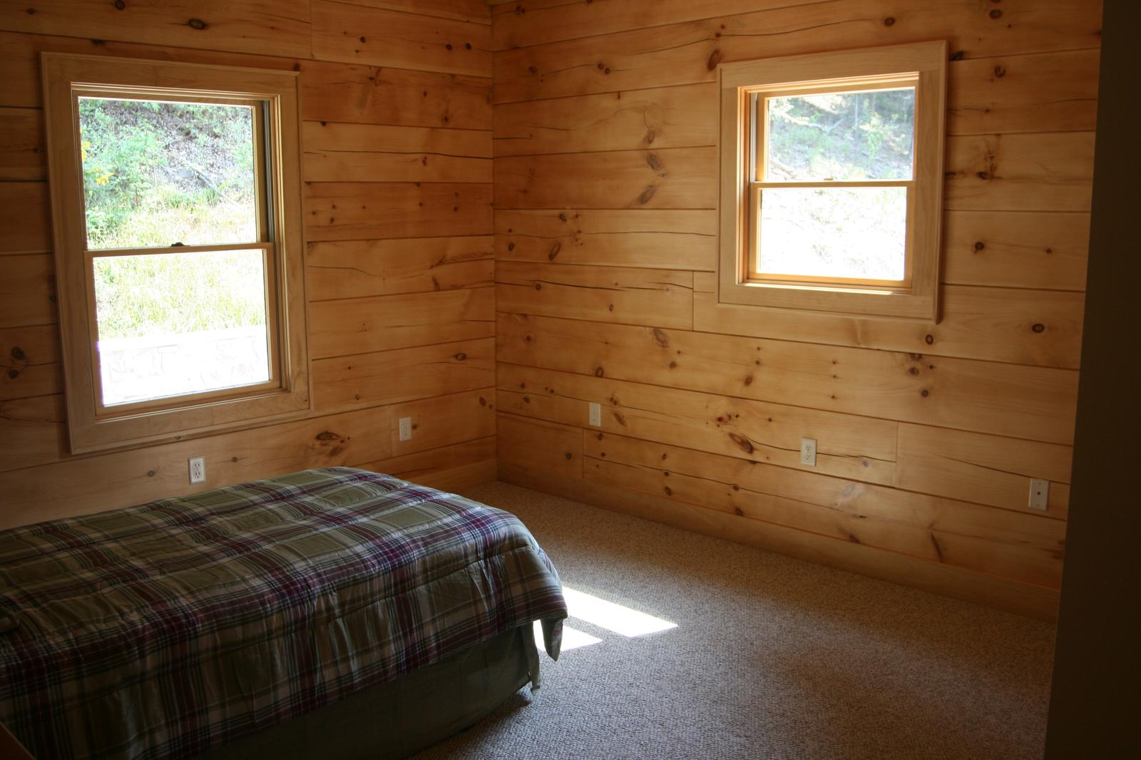 A bedroom with board walls and two windows, a plaid cover on the bed and light falling across the carpet