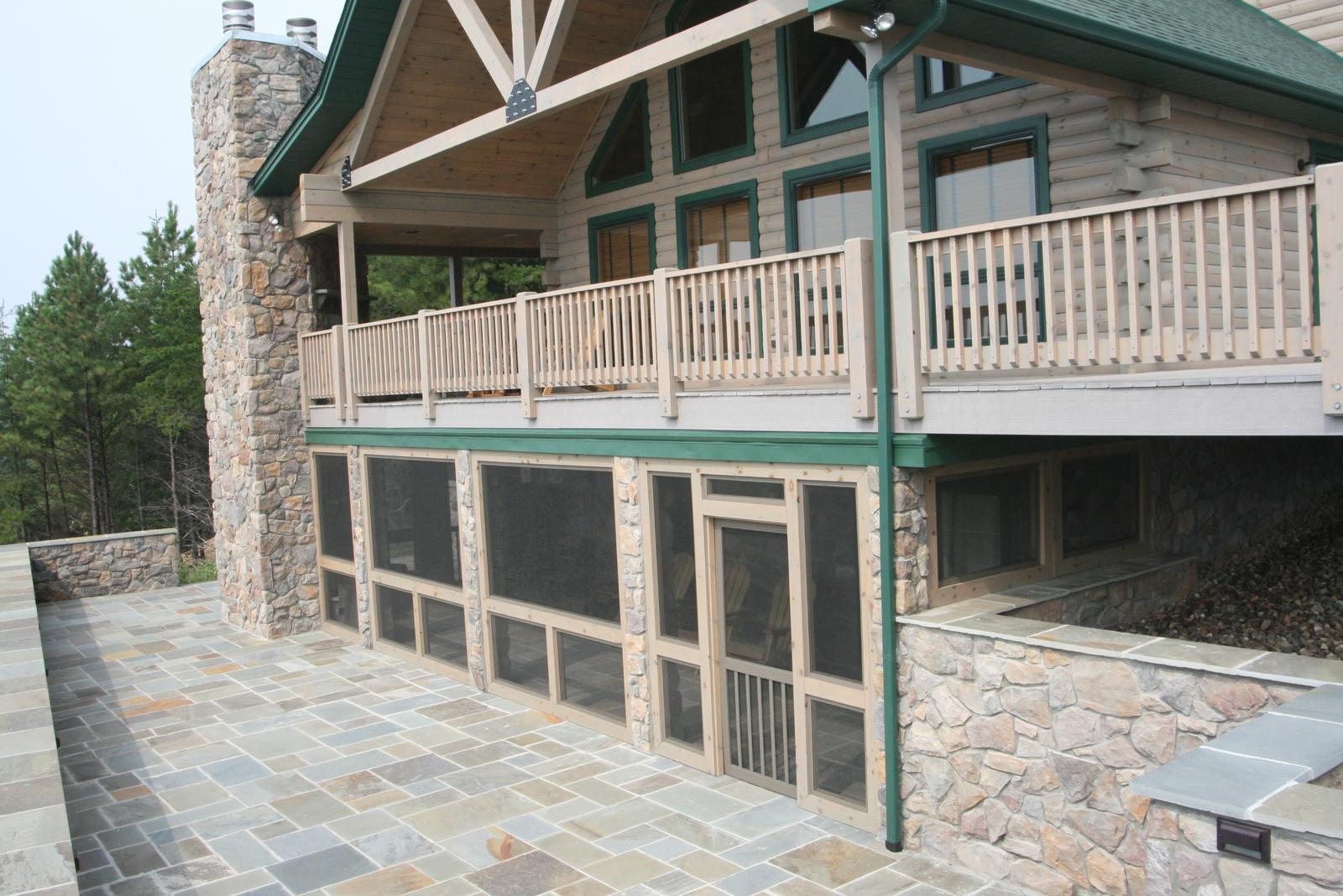 A screened lower level under a railed deck, stone columns and chimney rising past it above a flagstone terrace