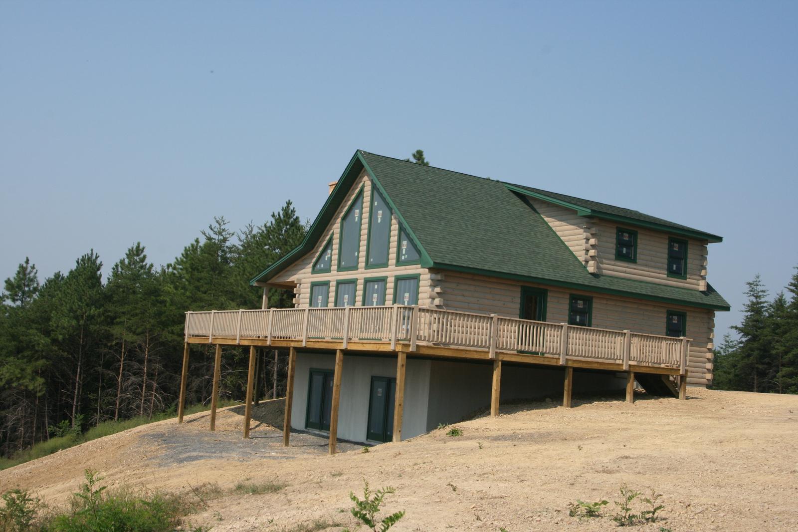 A finished log home with a green roof and a full-width deck on posts, standing on freshly graded ground
