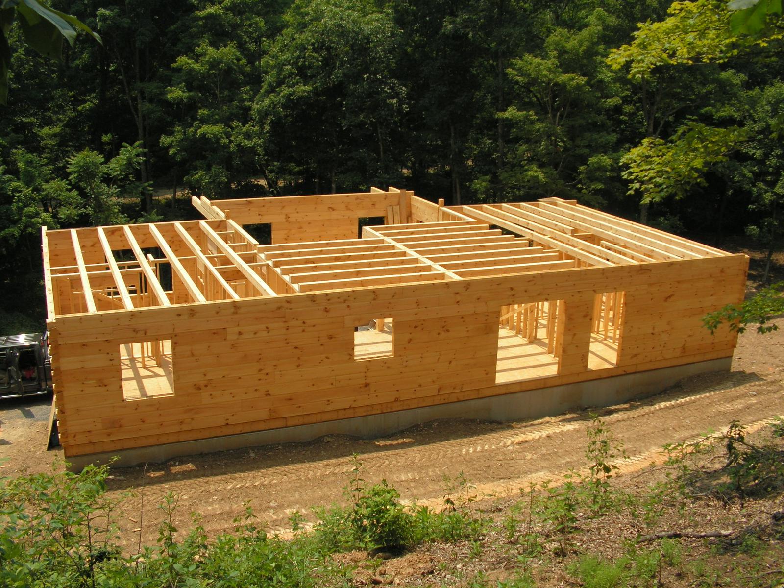 Log walls stacked to first-floor height with floor joists laid across, seen from above on a cleared slope