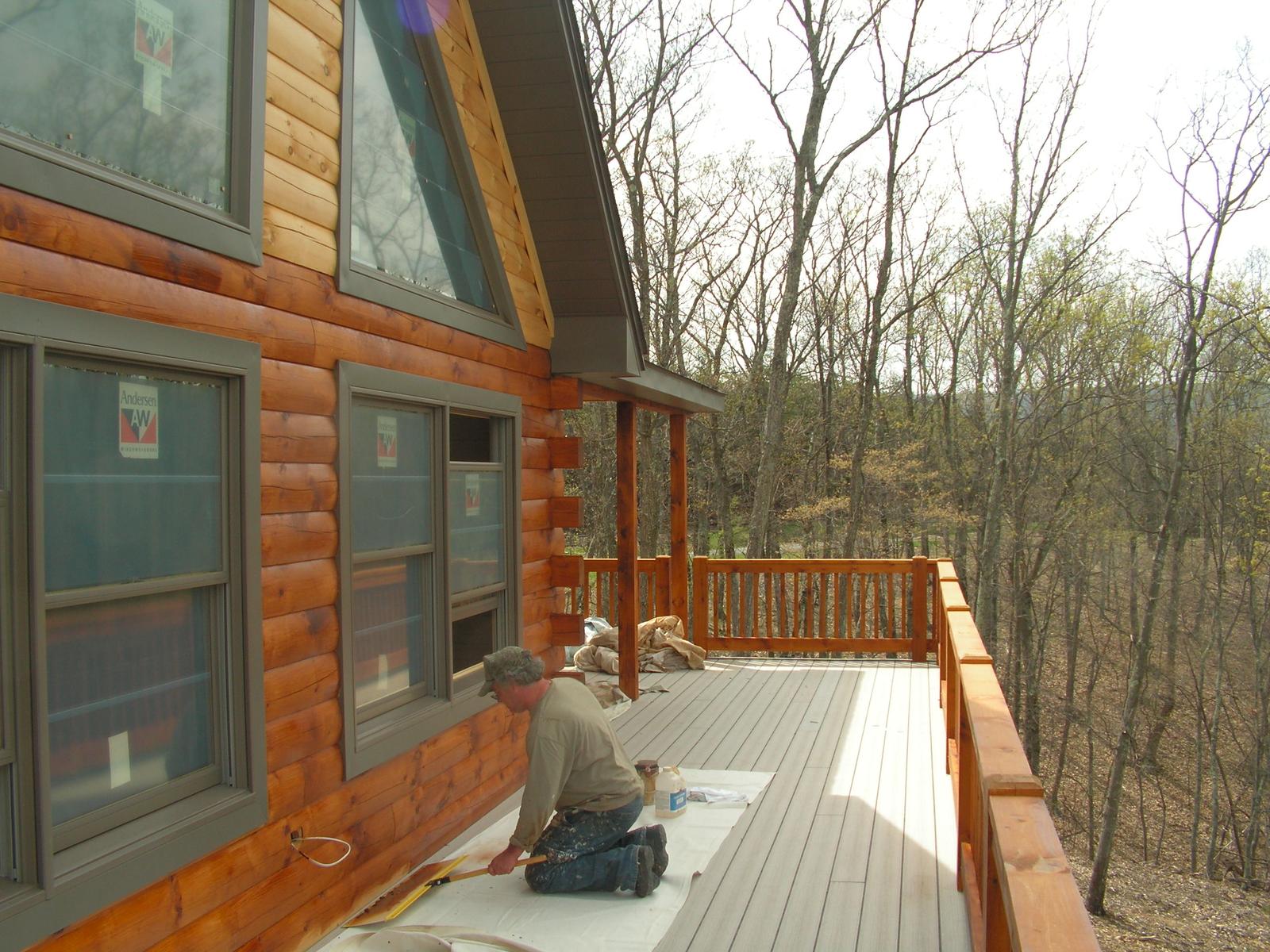 A worker kneeling on new decking beside a log wall, tools laid out beside him and bare woods beyond