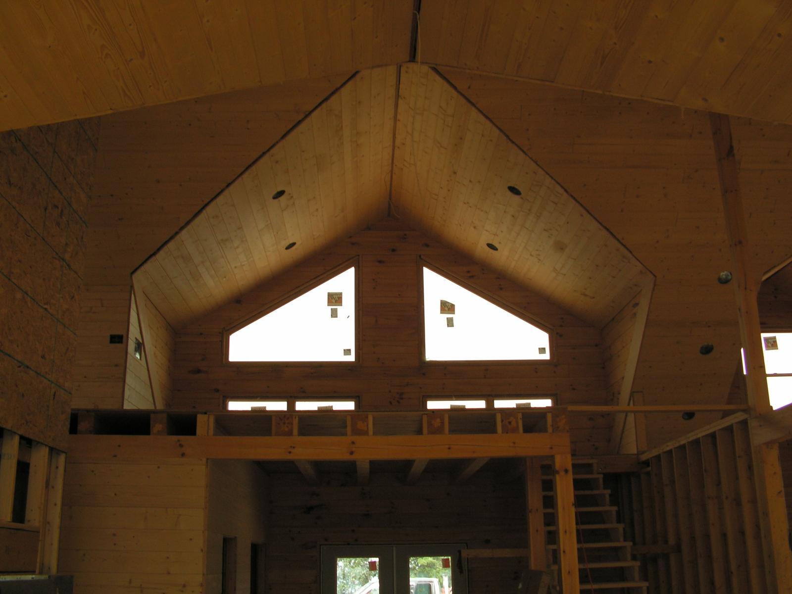 A vaulted board ceiling over an open room, two triangular gable windows above and a loft rail across the far end