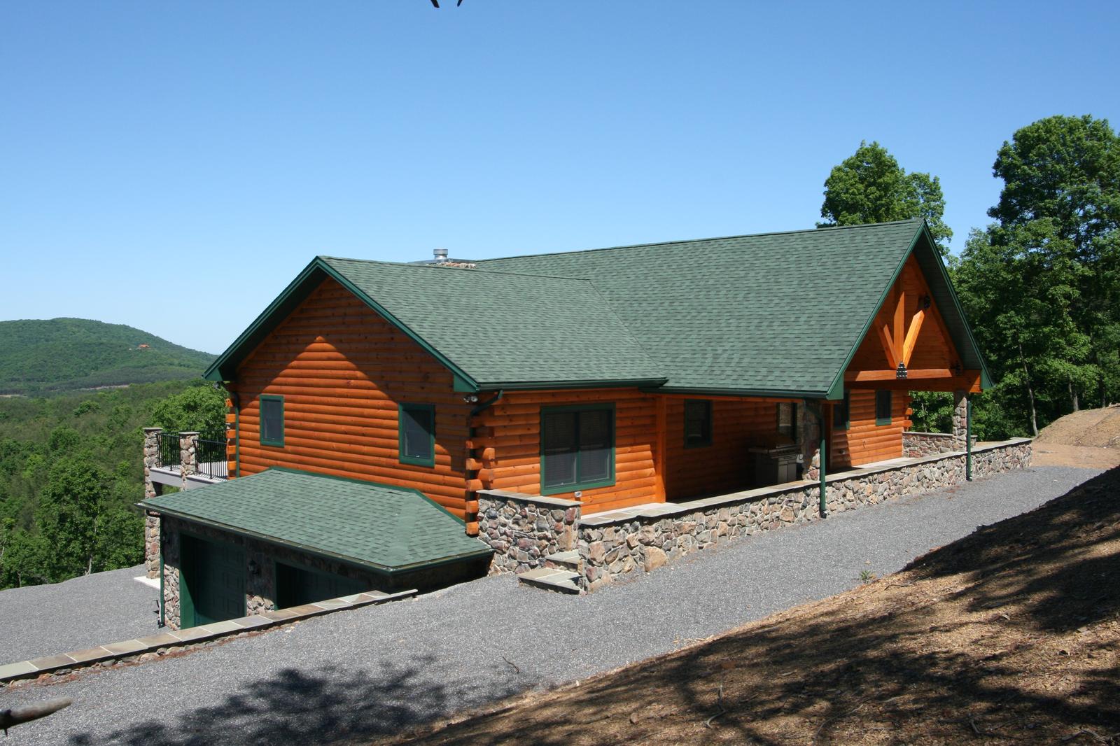A log home along its length from the drive, green roof over round log walls, stone retaining wall stepping down to the gravel, mountains behind