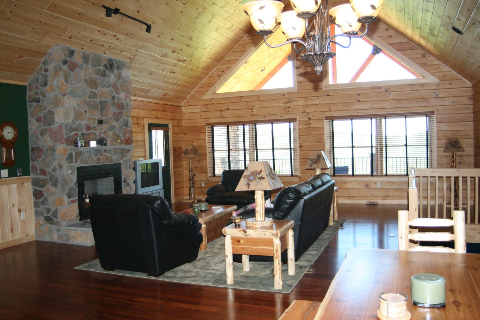 A great room under a vaulted board ceiling, a stone fireplace with a wood mantel at one end and triangular gable windows above, leather seating on a wood floor