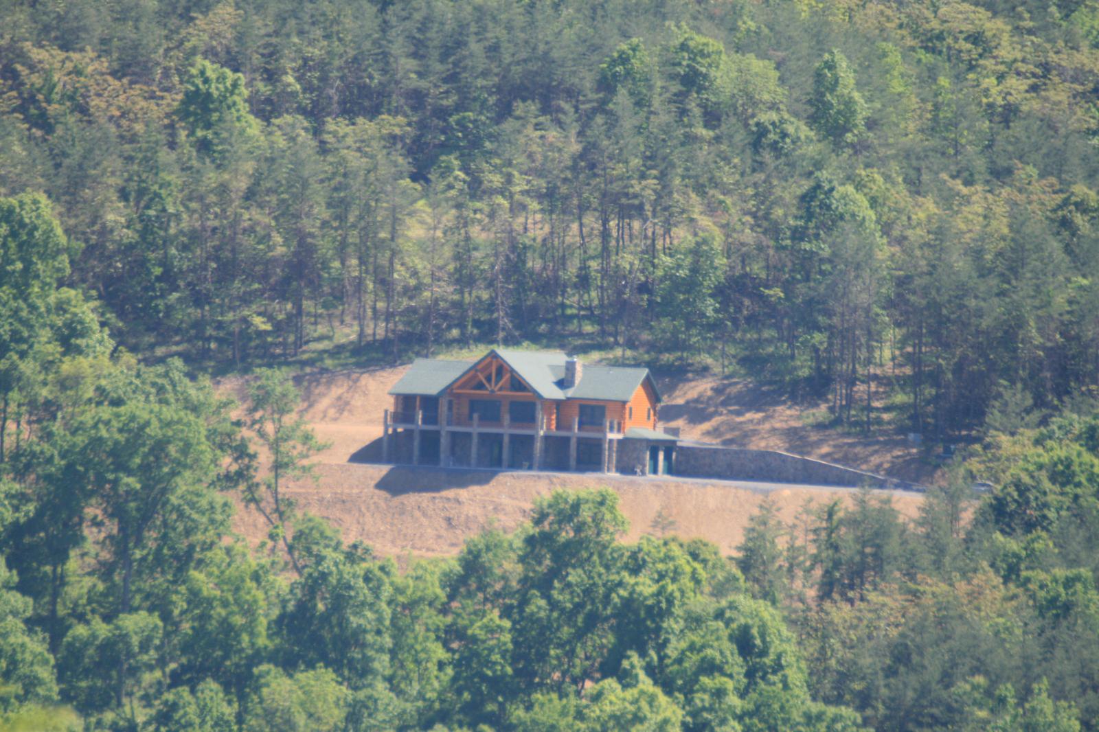 A log home alone in a cleared pocket, seen from across a wooded slope with trees closing in on every side