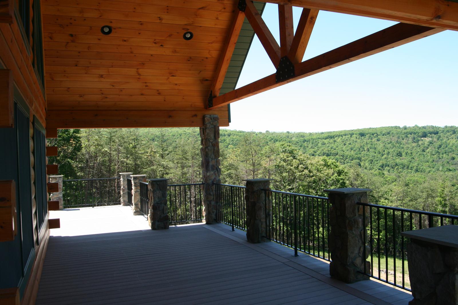 A covered porch open to a valley, exposed timber rafters and a truss overhead, stone columns and black railing across the view