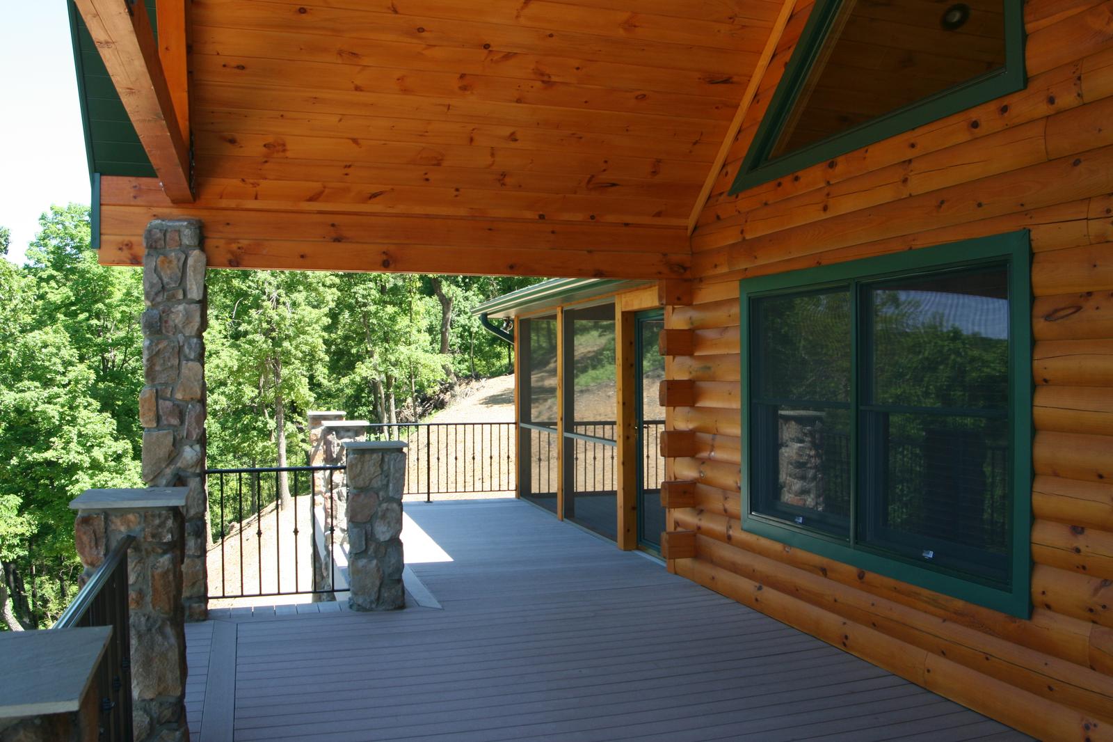 A covered deck running along log walls with green-trimmed windows, stone columns carrying the roof, woods close on the open side