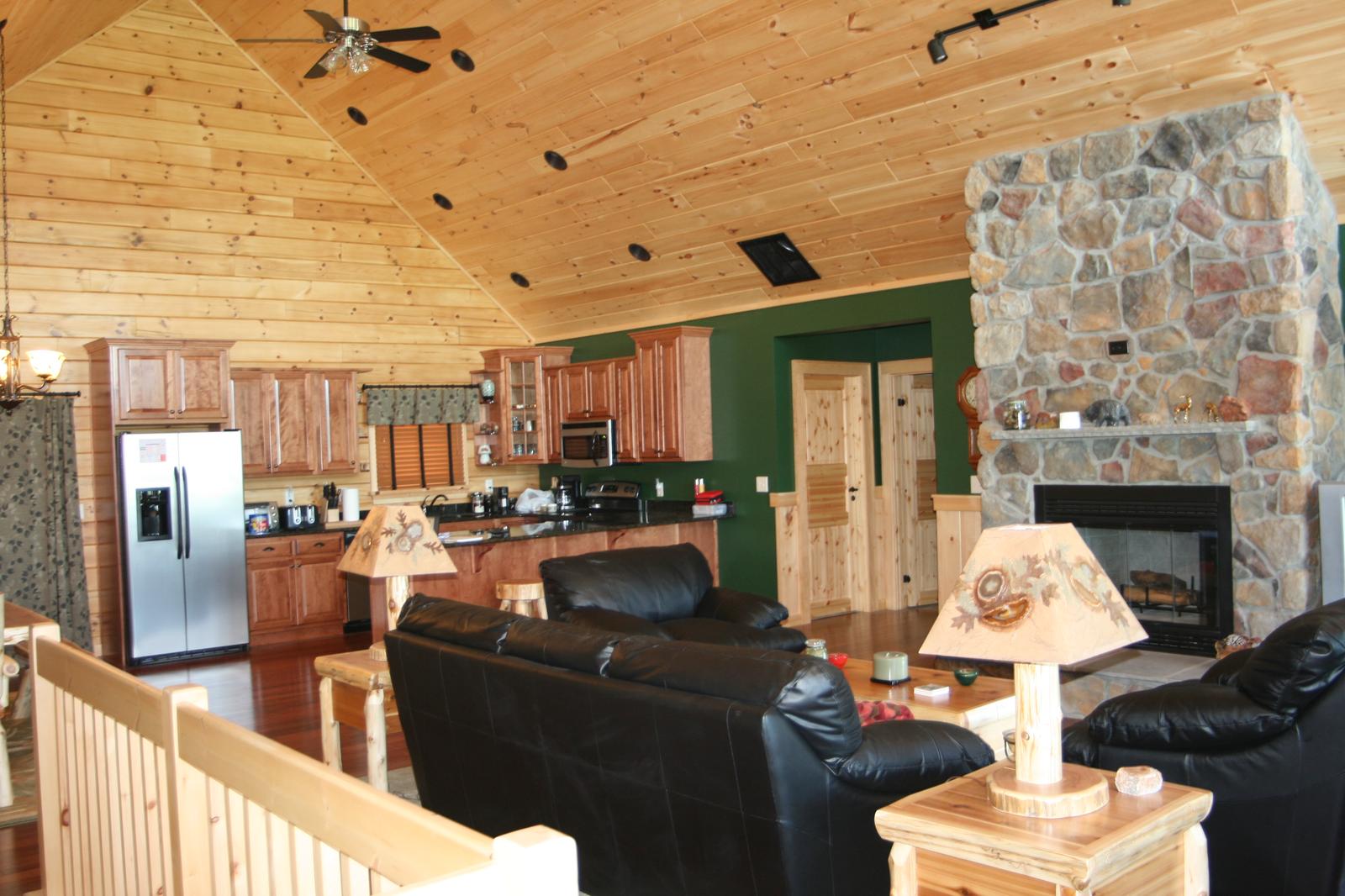 A great room and kitchen seen across a log stair railing, vaulted board ceiling with a fan, stone fireplace to one side and a dark green painted wall behind the cabinets