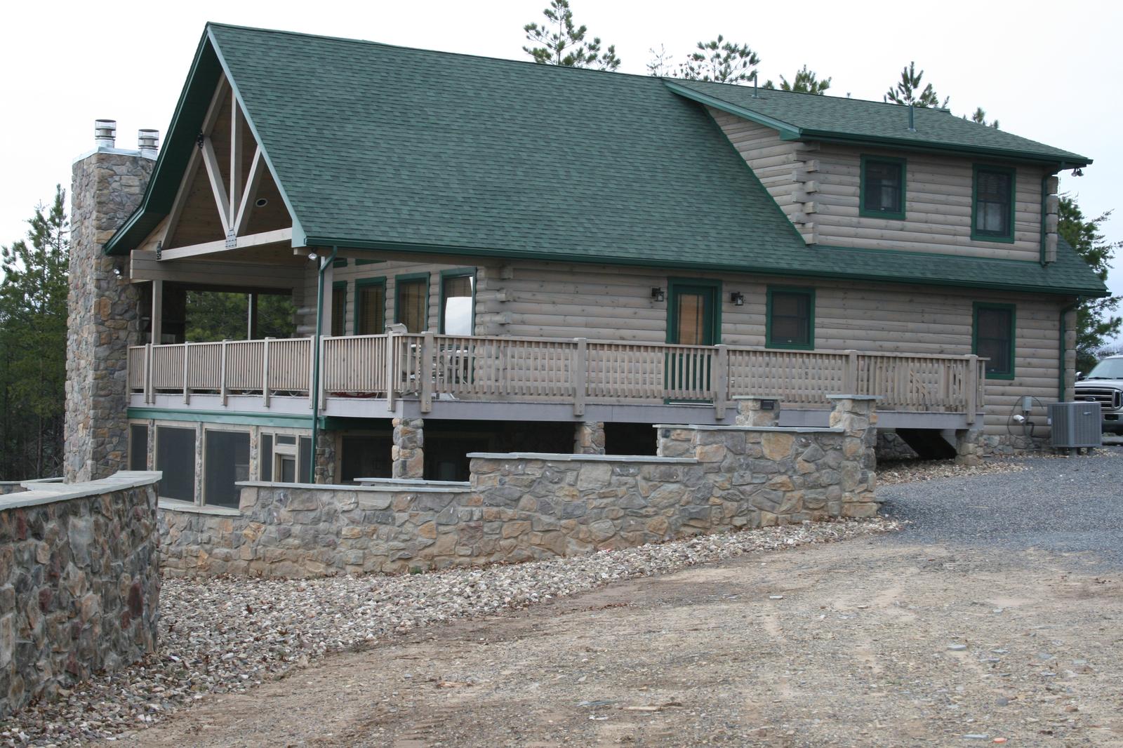 A log home from the gravel drive in winter, stone chimney and gable truss above a railed upper deck, the lower level faced in stone