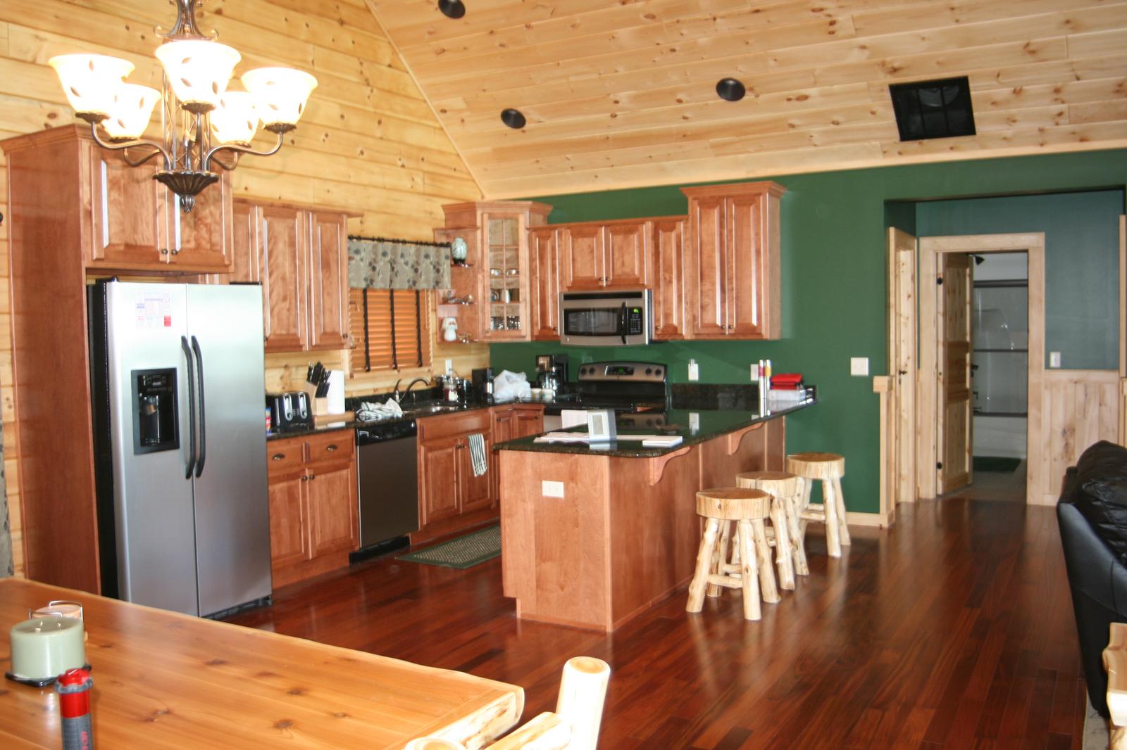 A kitchen with wood cabinets to the ceiling, stainless refrigerator and range, an island with log-post stools, a chandelier above and a dark green wall beyond