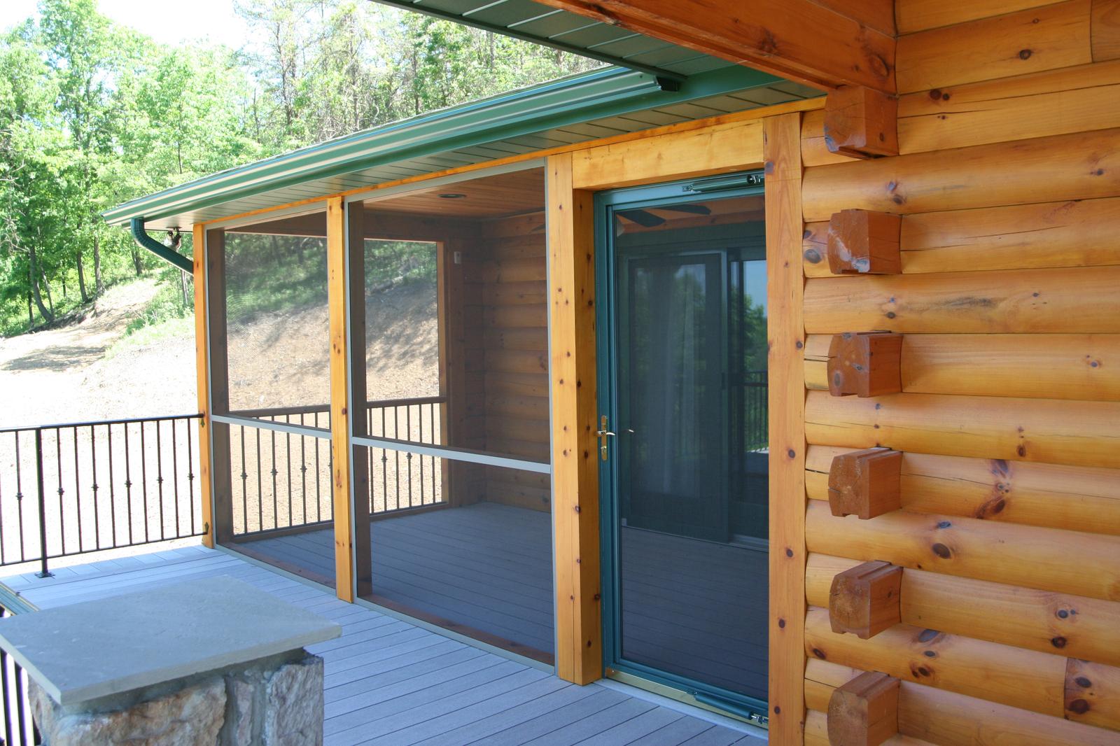 A screened section of porch beside a glazed door, heavy round log walls with projecting log ends, black railing and a stone pier at the corner