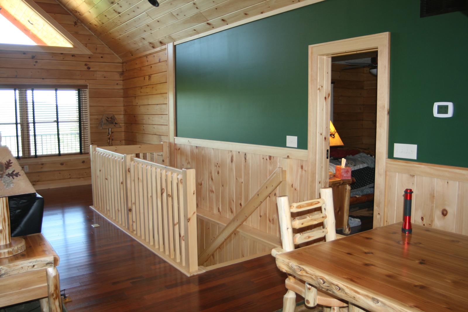 A room with one wall painted dark green above a vertical board wainscot, a log railing at the stair opening and log-frame chairs at a wood table