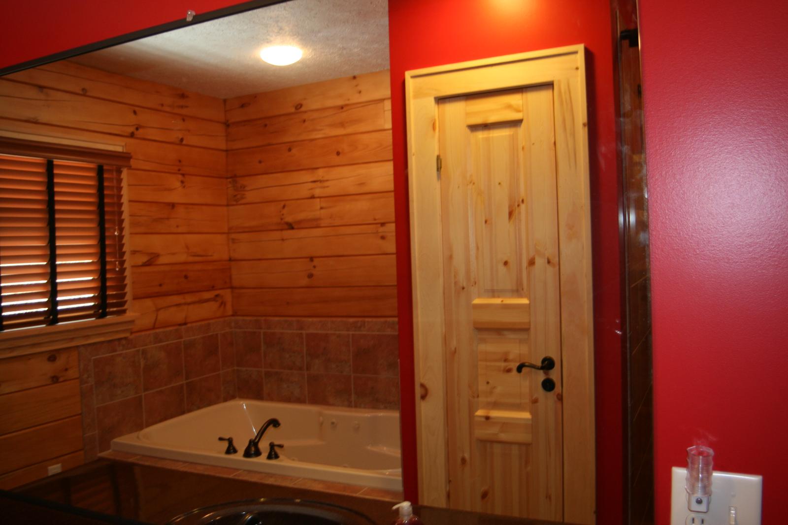 A corner tub set into a tiled surround below a shuttered window, board walls, a plank door and a red painted wall