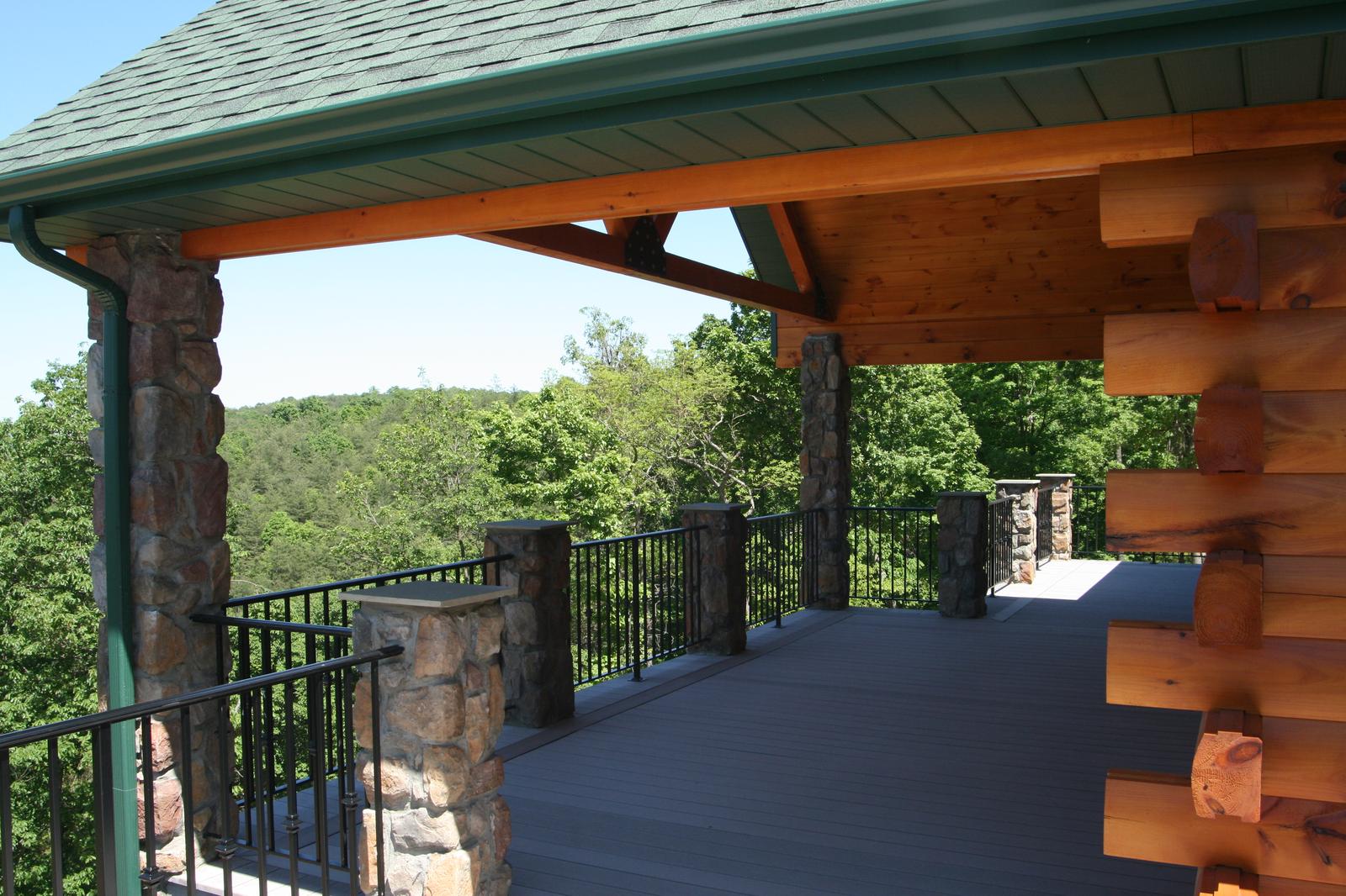 A covered deck under a green roof, log posts and log walls beside a black metal railing, wooded ridges filling the opening