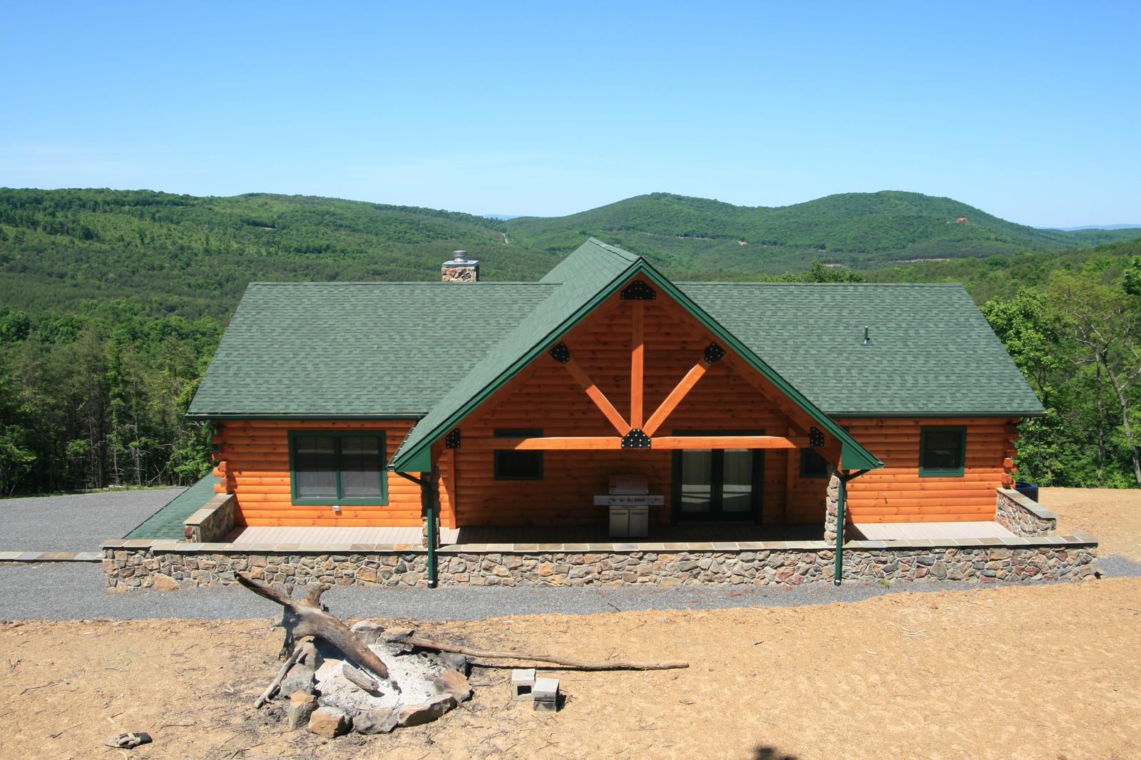 A log home square on, sunburst timber ornament centered in the gable above the entry, stone base and green roof, hills stacked behind