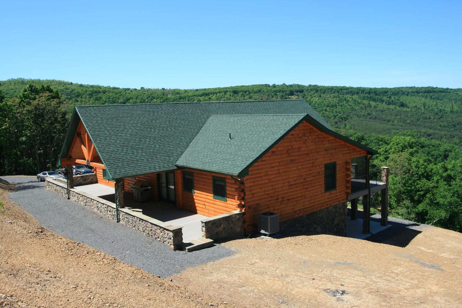 A log home from above and to one side, green roof and stone base, a graded drive curving in and the valley beyond