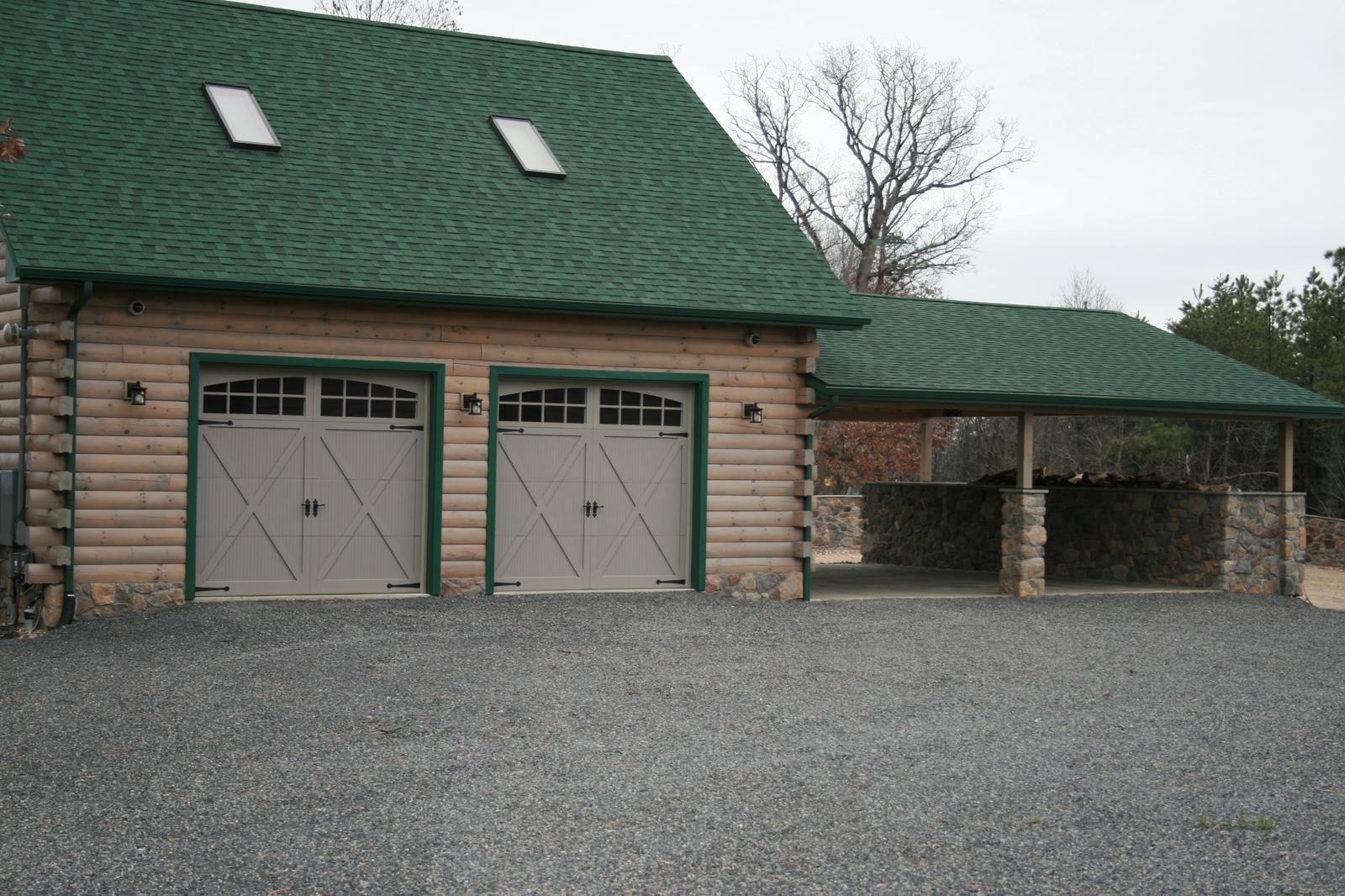 A detached garage with two carriage-style doors under a green roof with skylights, a covered bay to one side standing on stone piers, gravel apron in front