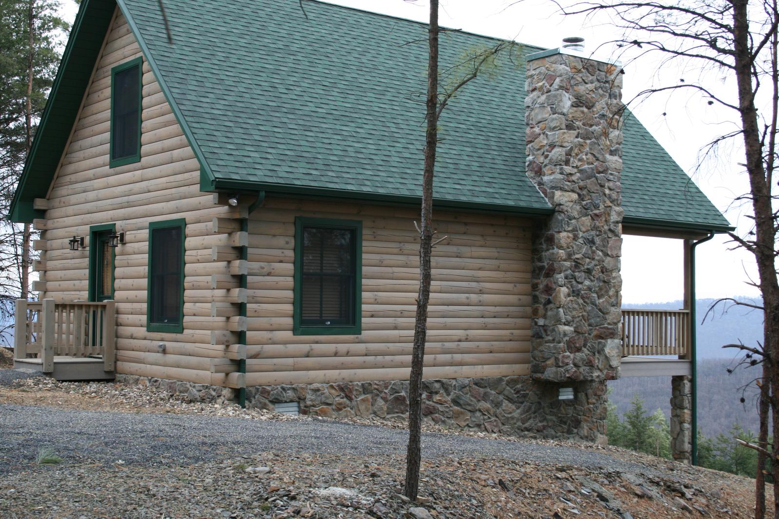 A log home from the drive, green roof and green-trimmed windows above a stone foundation skirt, a tall stone chimney on the near gable