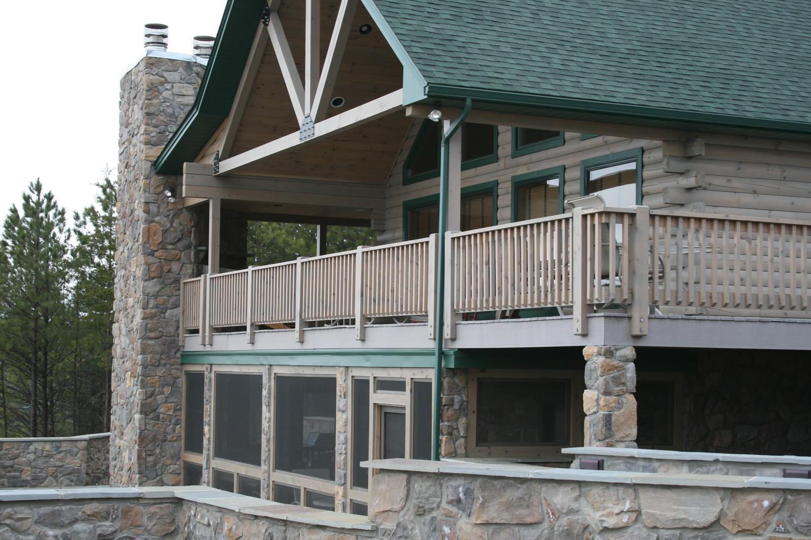 A log home seen from below the retaining wall, a stone chimney running the full height beside a timber truss in the gable, screened lower level under the deck