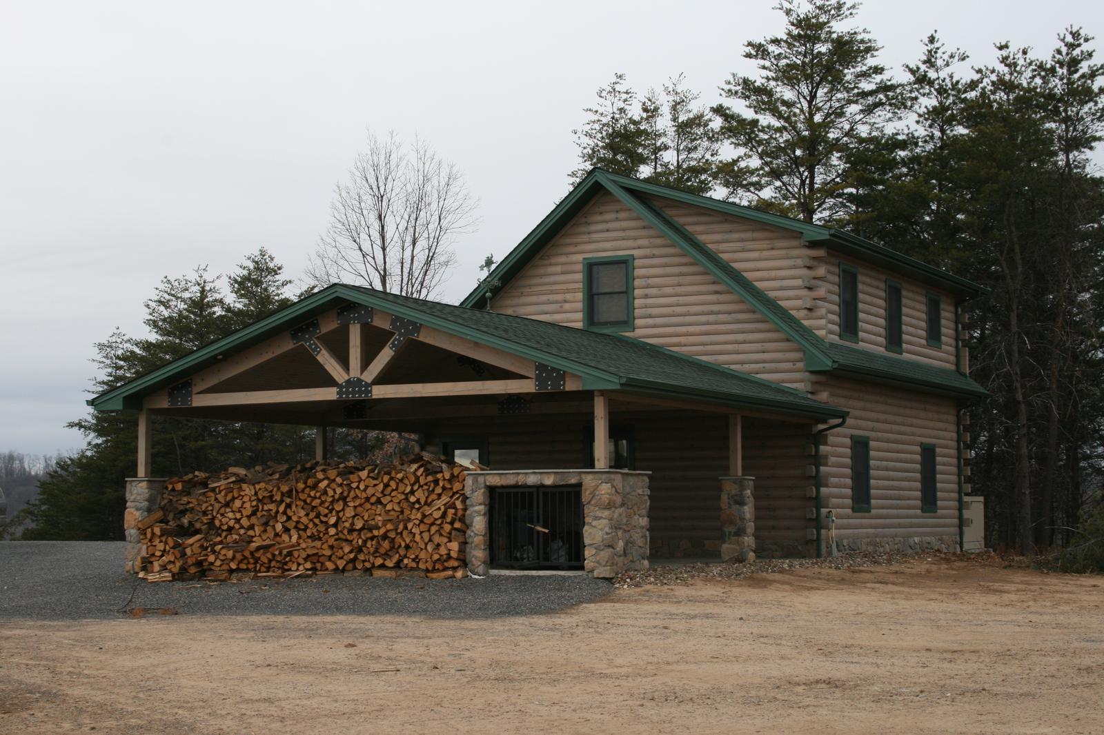 A covered bay stacked with split firewood beside a garage, a timber truss in its gable, bare ground and gravel in front