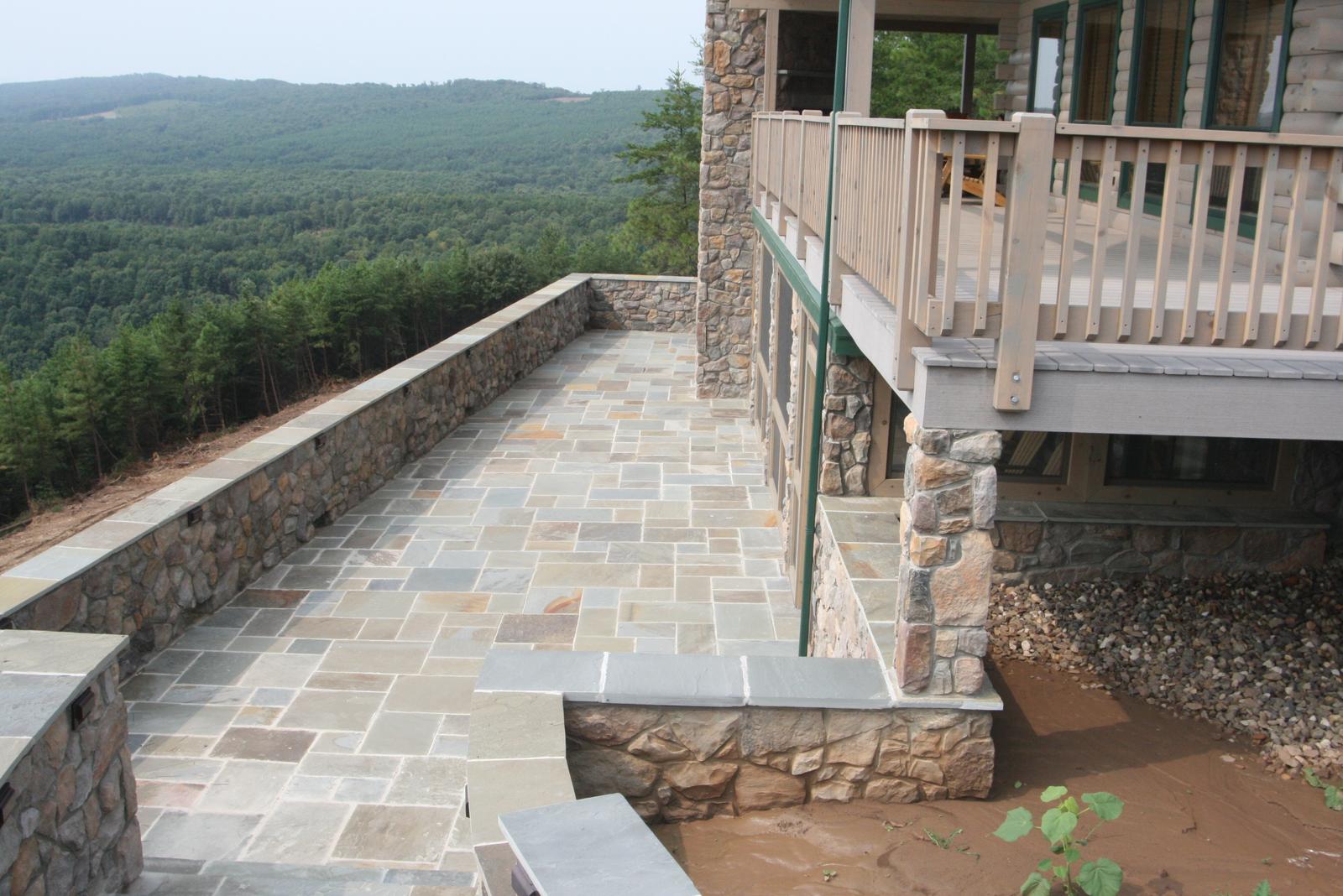 A flagstone terrace behind a low stone wall running the length of the house, the deck above carried on stone columns, wooded ridges falling away beyond