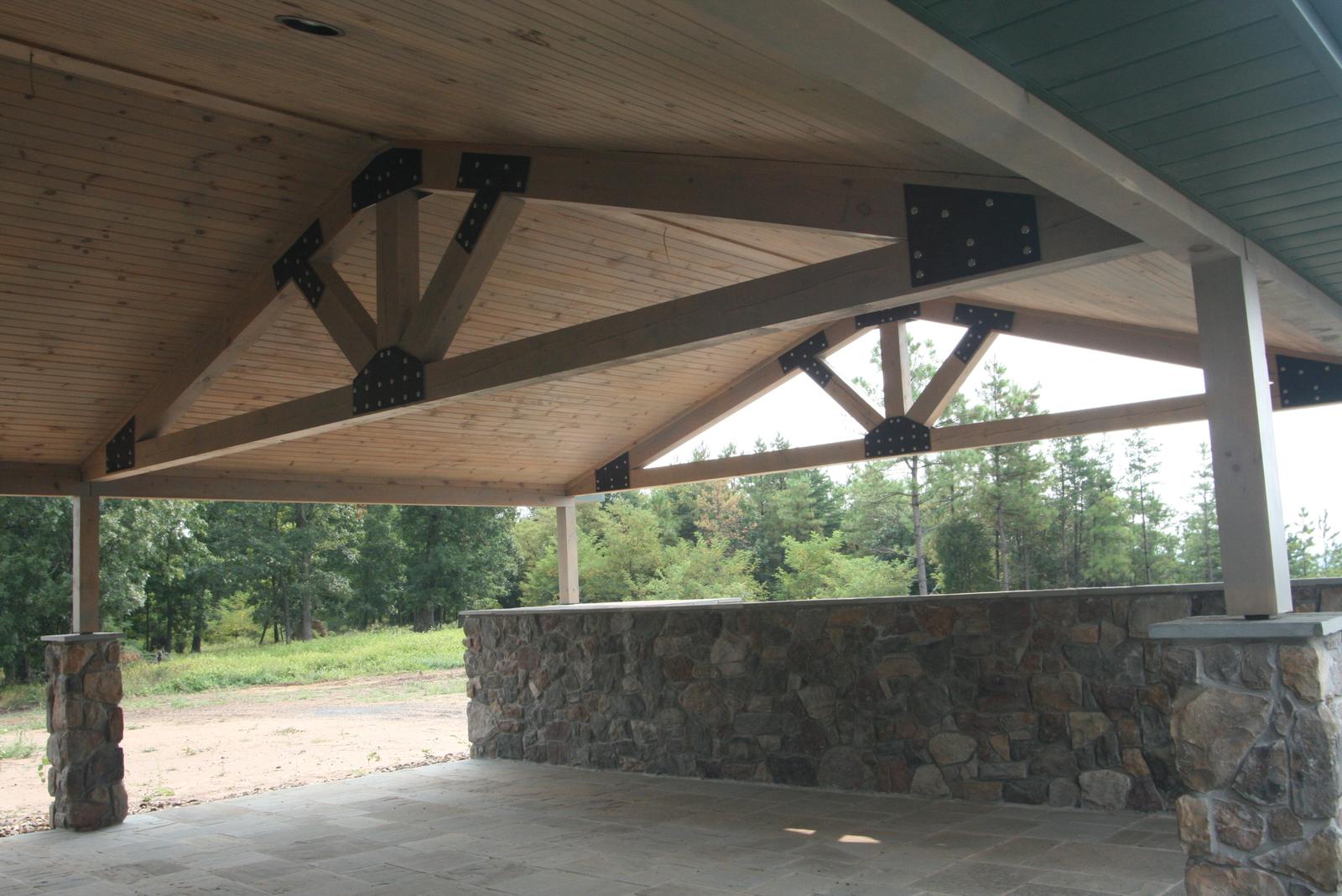The underside of a covered porch roof, exposed timber trusses joined with black steel plates and bolts against a board ceiling, stone knee-wall and columns below