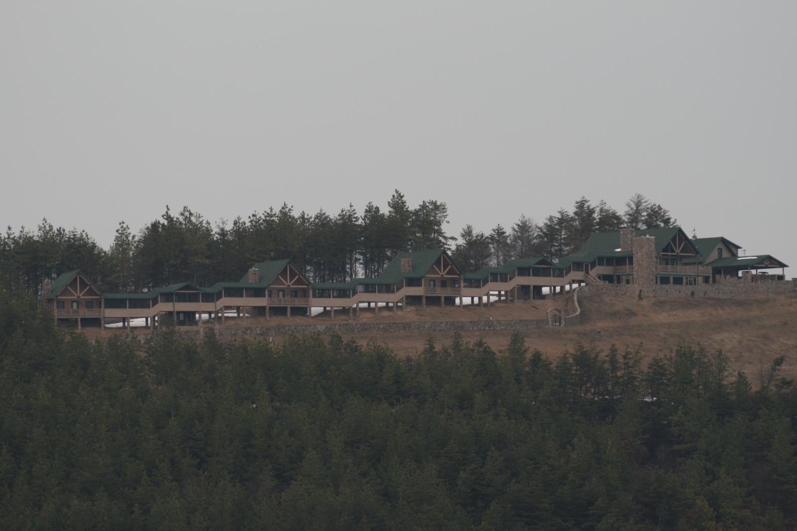 A row of houses spread along a ridge in haze, seen from far below across a stand of evergreens