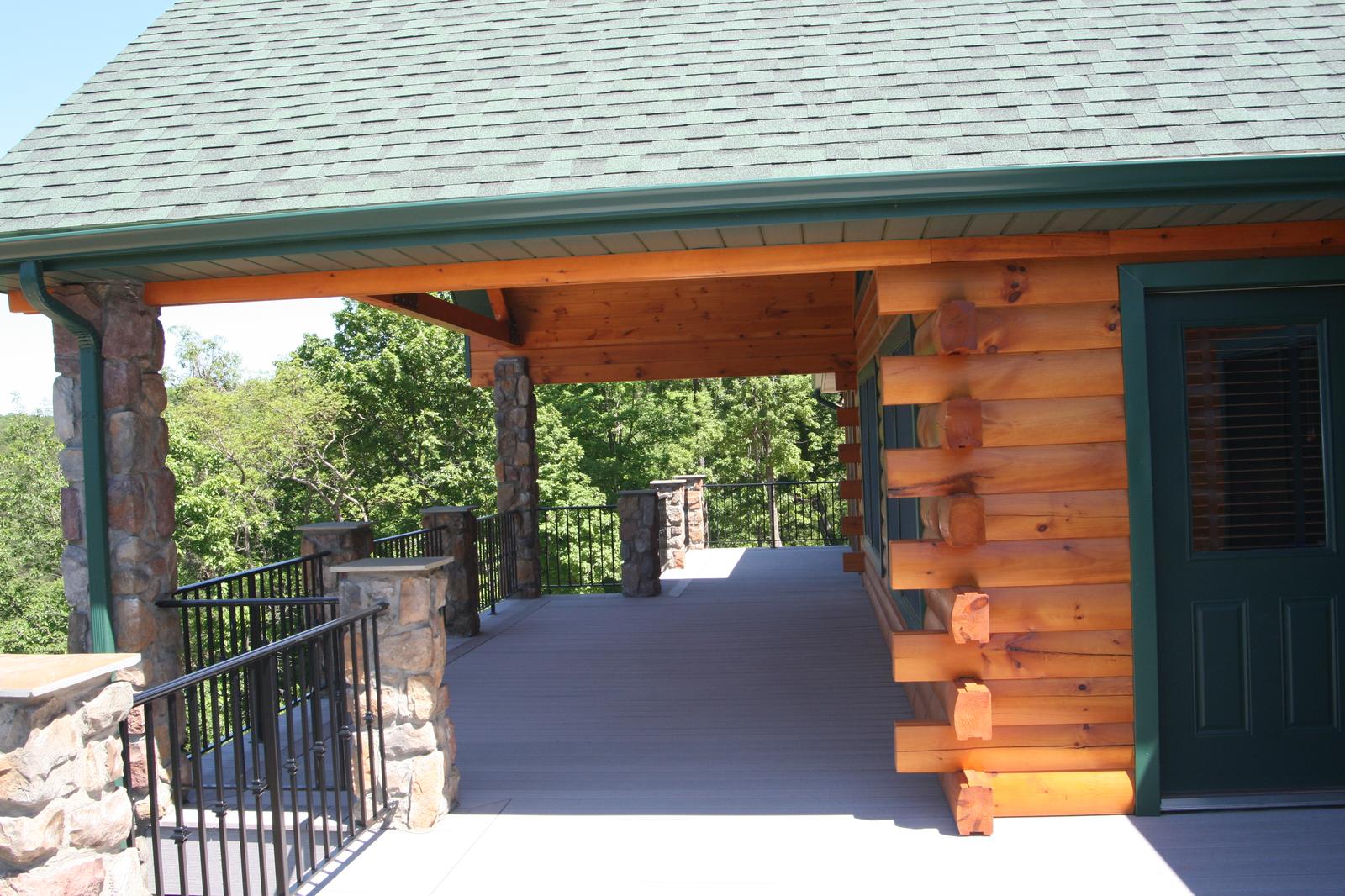 A covered walkway between stone columns along a round-log wall, a green-trimmed door at the end and the ridge beyond the railing
