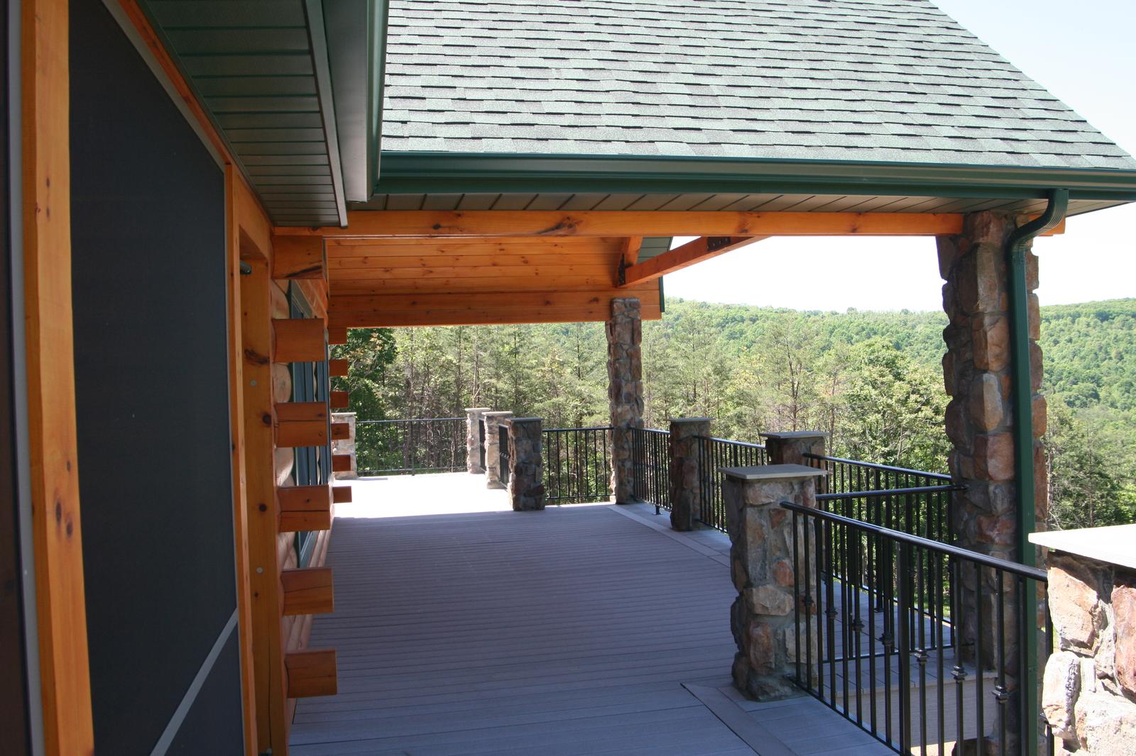 A covered porch running the length of a log wall, its roof carried on timber posts and stone columns, valley beyond the black railing