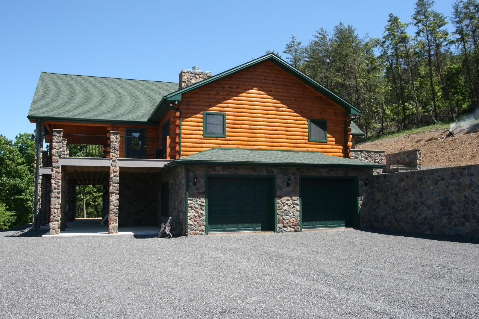A log home from the gravel drive with a two-bay garage set into its stone lower level, green roof above, cut bank and retaining wall behind