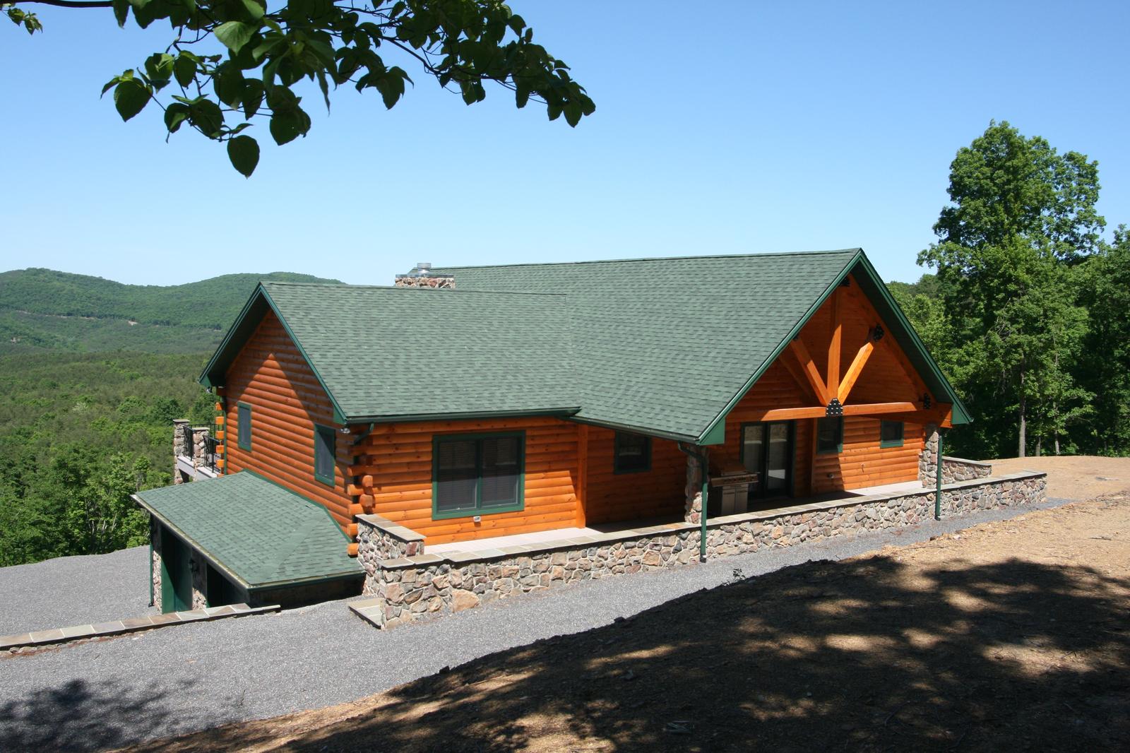 A log home on a ridge with a sunburst timber ornament in the gable, green roof and stone base, cleared ground falling away in front