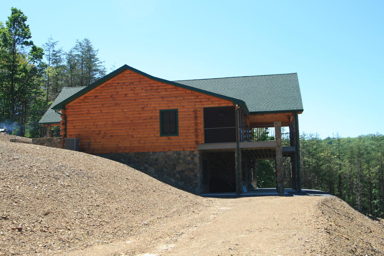 A log home standing on a raw bank, green roof over log walls and an open walkout level below, bare graded earth in front