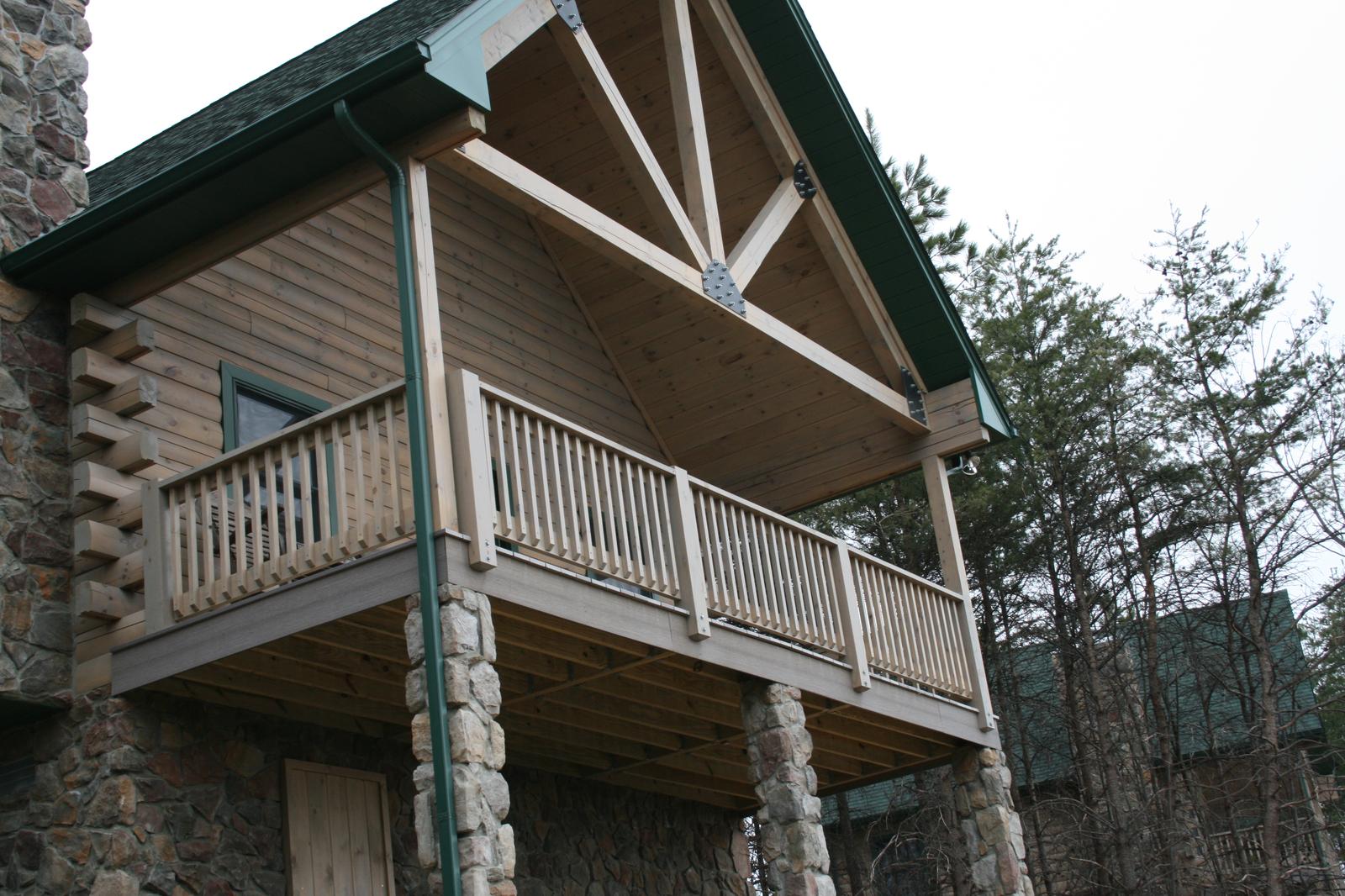 A deck corner seen from below, log walls and a timber truss in the gable above, the deck carried on stone columns
