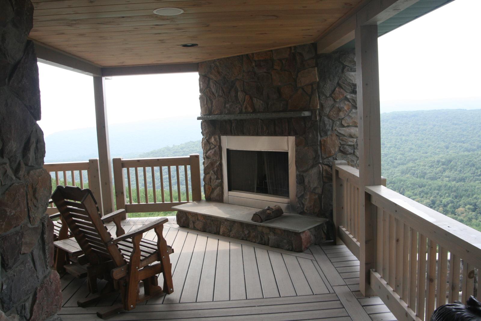 An outdoor stone fireplace built into the end of a covered porch, two wooden rocking chairs drawn up to it, the valley beyond the railing