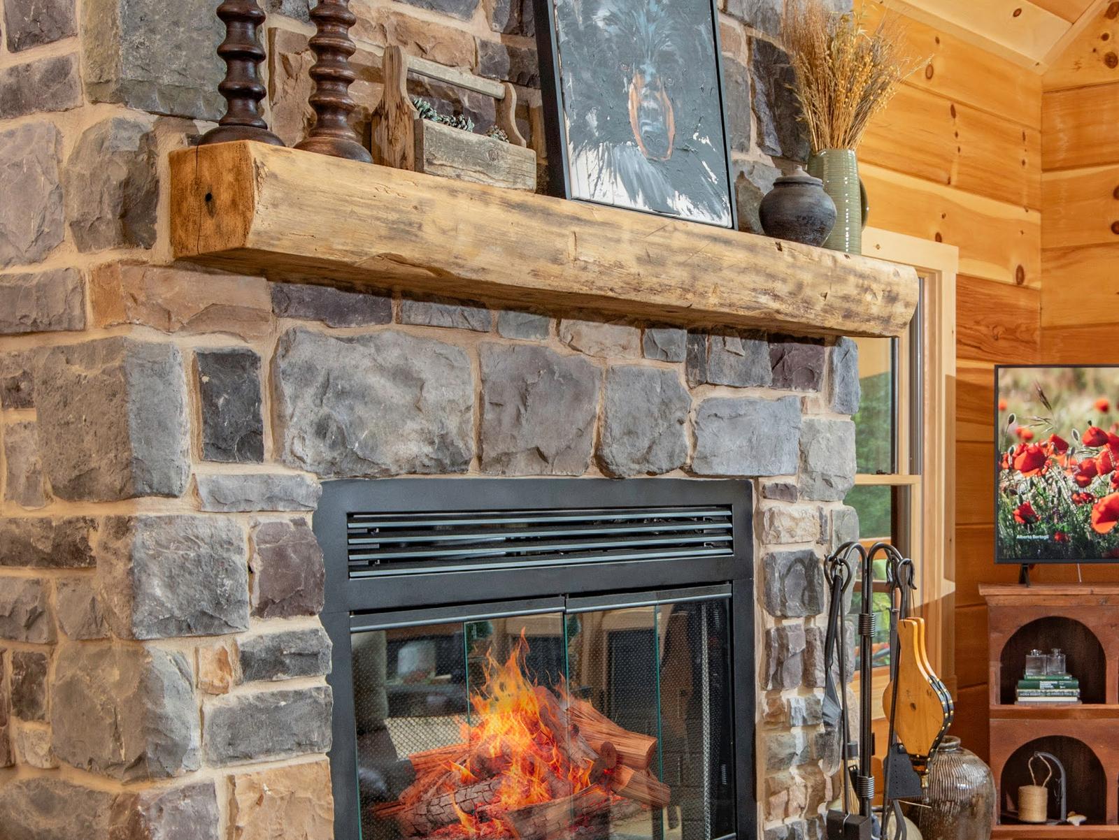 Close view of a hand-hewn timber mantel set into a rough fieldstone chimney breast, adze marks and checking visible along the beam, a lit firebox below