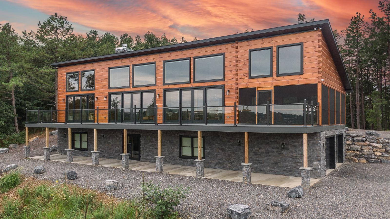 A modern log home at sunset, with a long glass-railed deck over a stone walkout and an orange sky above the roofline
